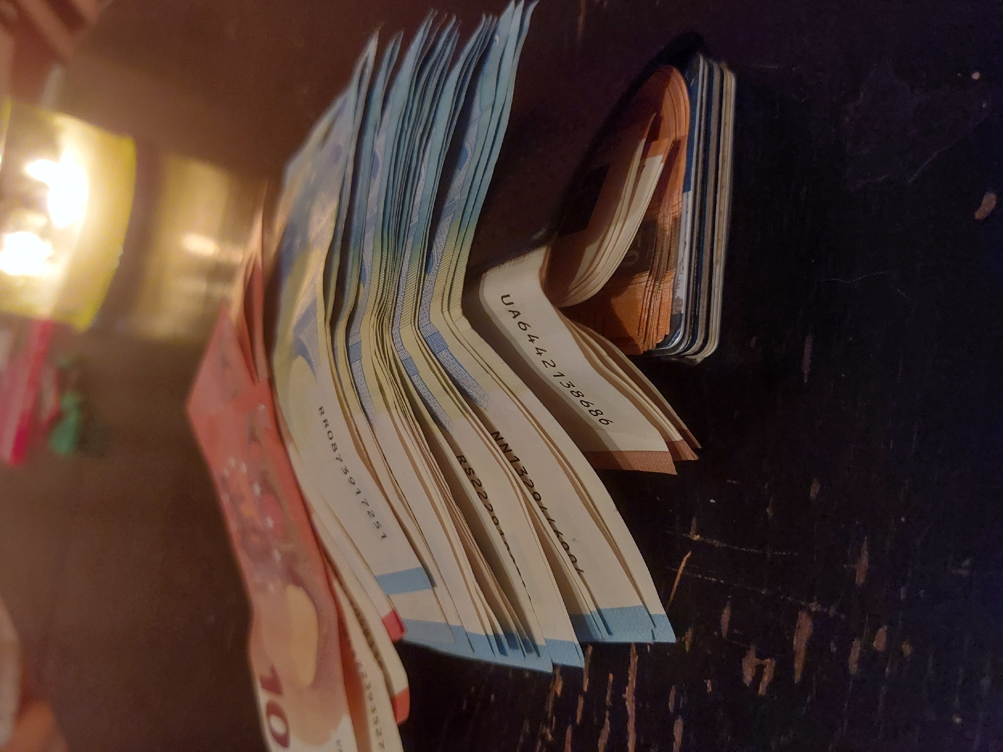 The image shows a stack of euro banknotes, arranged in an upright, slightly curved position on a dark, possibly wooden surface. The banknotes appear to be a mix of denominations, with some identifiable as 10 euro notes (reddish-orange) and others as 20 euro notes (blue). Serial numbers are visible on some of the notes. A blurry light source is present in the background. The banknotes are the primary subject of the image. The location (Tsarevo, Bulgaria) is not visually discernible from the image.