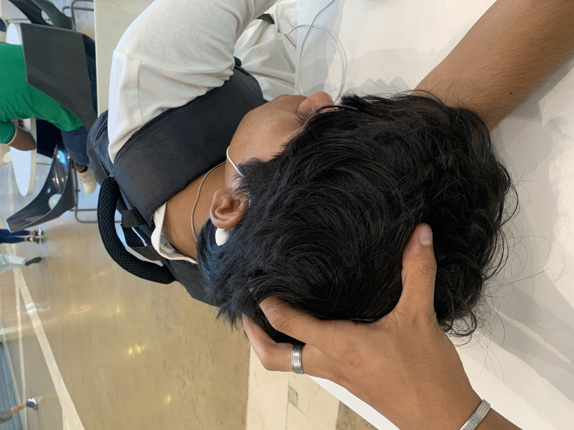 This media file depicts an intimate moment between two individuals in what appears to be a bright, indoor public space in Pampatar, Venezuela. The main subject, viewed from above, is a person resting their head on a pristine white surface, likely a table. This individual has dark, wavy hair and is wearing a white long-sleeved shirt over which a dark backpack with visible mesh straps is donned. A silver chain necklace is visible around their neck, and a thin white cord, possibly from an earbud or a face mask, is near their ear.
Another person's hand is gently placed on the head of the resting individual, seemingly cradling or stroking their hair, conveying a sense of comfort or care. This hand features a silver ring on the ring finger and a silver bracelet with a Greek key design on the wrist.
The background suggests a spacious public environment, possibly a food court, waiting area, or a large hall. The floor is light-colored, possibly polished concrete or large tiles, and features distinct white lines, which could be part of a court or pathway markings. Other partially visible individuals, chairs, and tables are present in the background, including a person in a green shirt. The lighting is bright and even throughout the scene, indicative of either good artificial illumination or ample natural light. On the collar tag of the resting person, the text "30 OE" is partially visible. The overall mood is one of quiet solace amidst a public setting.