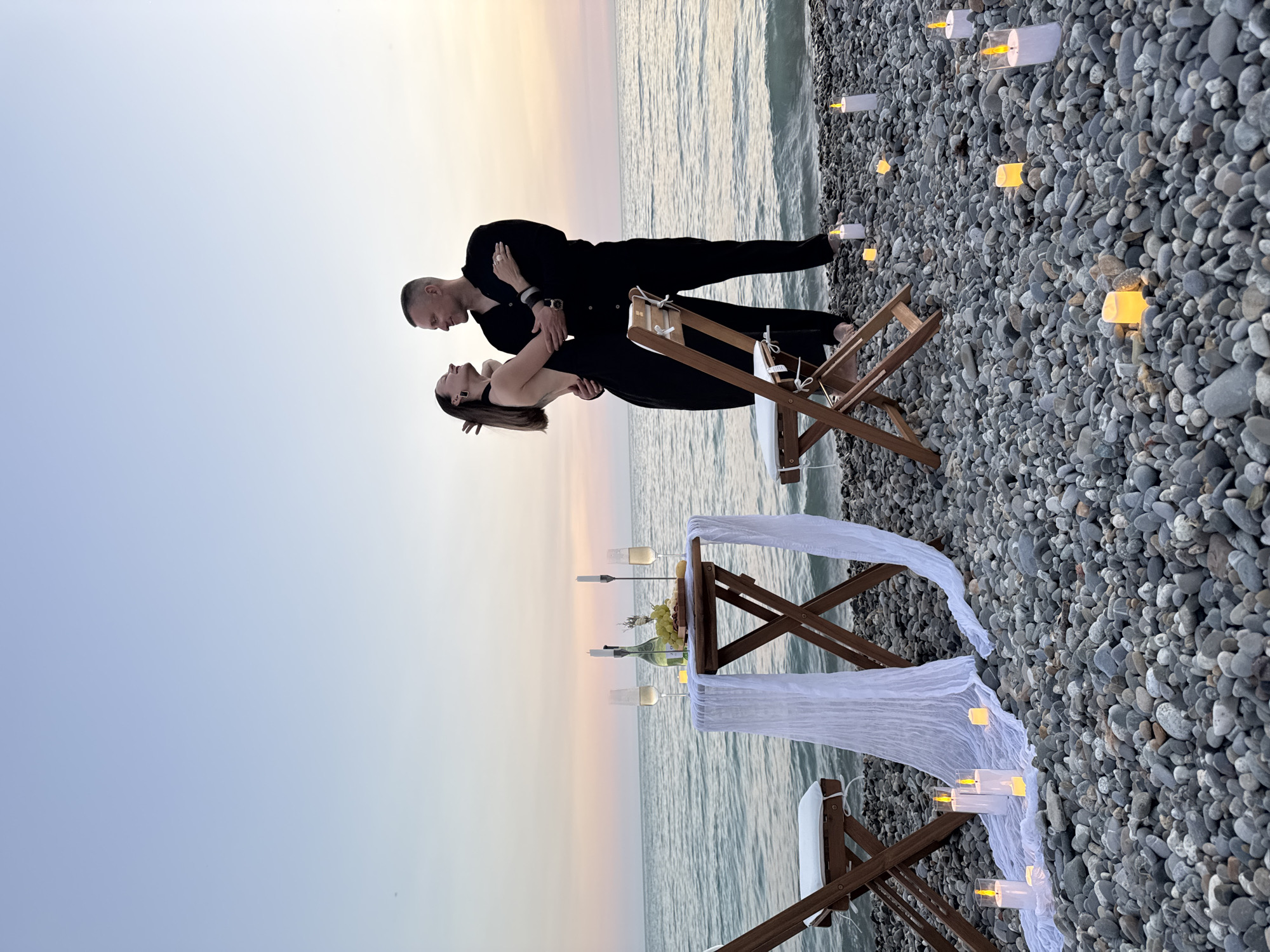 A couple in black clothing is standing on a pebble beach by the sea at dusk, with a table set for two with champagne, grapes, and candles. The scene is illuminated by the warm glow of numerous candles scattered across the beach. The water is calm, and the sky displays soft hues of orange and purple from the setting sun. The location is likely a secluded spot on the coast of Sochi, Russia, as suggested by the pebbled shore and the overall ambiance often associated with the Black Sea resorts during twilight. The couple appears to be in an intimate moment, with the man embracing the woman, who is looking up at him with a smile. The table is draped with white fabric, adding a touch of elegance to the romantic setup.