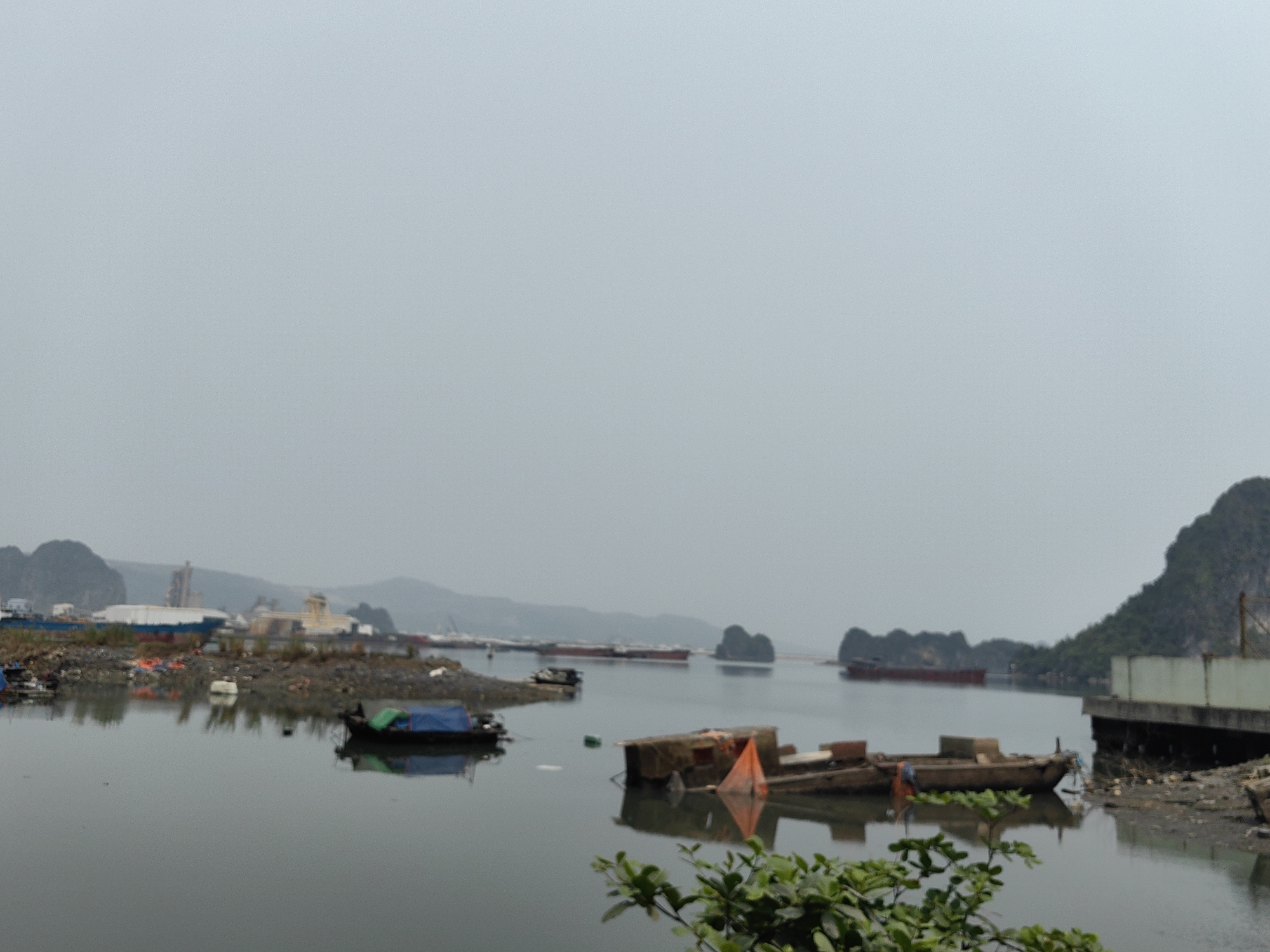 The image depicts a waterway scene in Quang Hanh Ward, Vietnam. Several small boats are docked along the shoreline. In the background, there are industrial structures and distant land formations. The sky is overcast, giving the scene a muted and gray tone. Vegetation is present in the foreground. The water appears calm and still, reflecting the surrounding landscape. Debris and trash are visible along the shoreline and in the water.