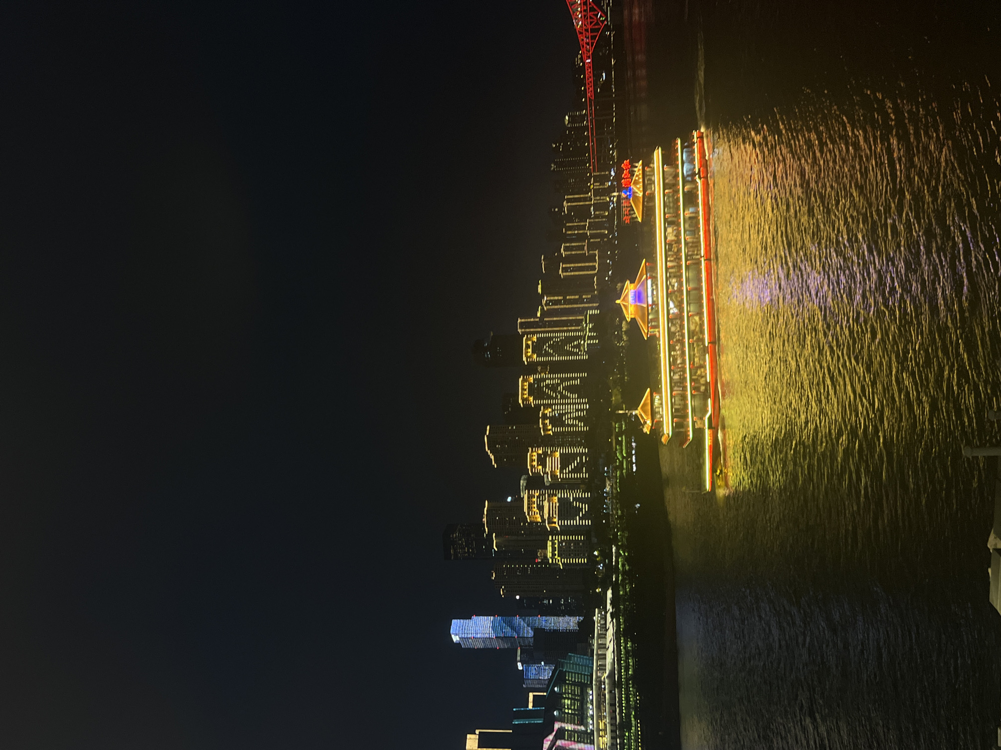 This is a nighttime photograph capturing the illuminated skyline of Yuzhong District, China, with a brightly lit tour boat in the foreground. The city's modern skyscrapers are adorned with golden lights, creating a vibrant contrast against the dark sky. A red illuminated bridge spans the river in the distance. The tour boat, a prominent feature, is decorated with colorful lights, reflecting a golden glow on the water's surface. The water itself appears dark, with ripples catching the ambient light. No people are clearly discernible due to the distance and lighting conditions.
