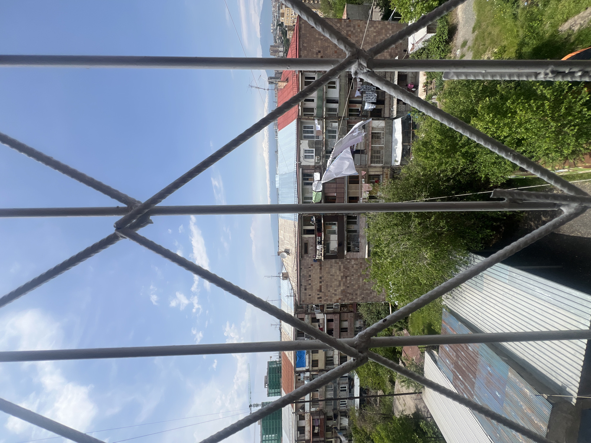 View from a balcony or window in Yerevan, Armenia, looking out over densely packed apartment buildings. A white flag or piece of fabric hangs from a clothesline strung between buildings. The sky is blue with scattered clouds. The foreground features a metal railing or scaffolding. Green trees are visible. In the distance are cityscapes and distant mountains.