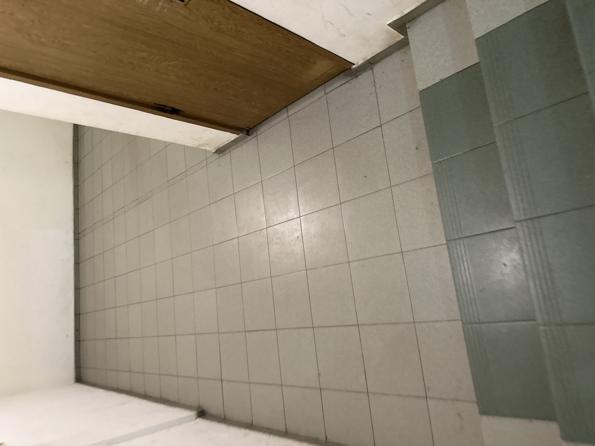 The image shows a section of tiled flooring and a lower portion of a door, likely in an interior space. The tiles are square and light-colored, with some green tiled sections at the top right of the frame. The door is wooden, and appears to be angled. A white wall is visible at the lower left of the frame. The location is unknown and not indicated within the image. The scene is a simple architectural detail.