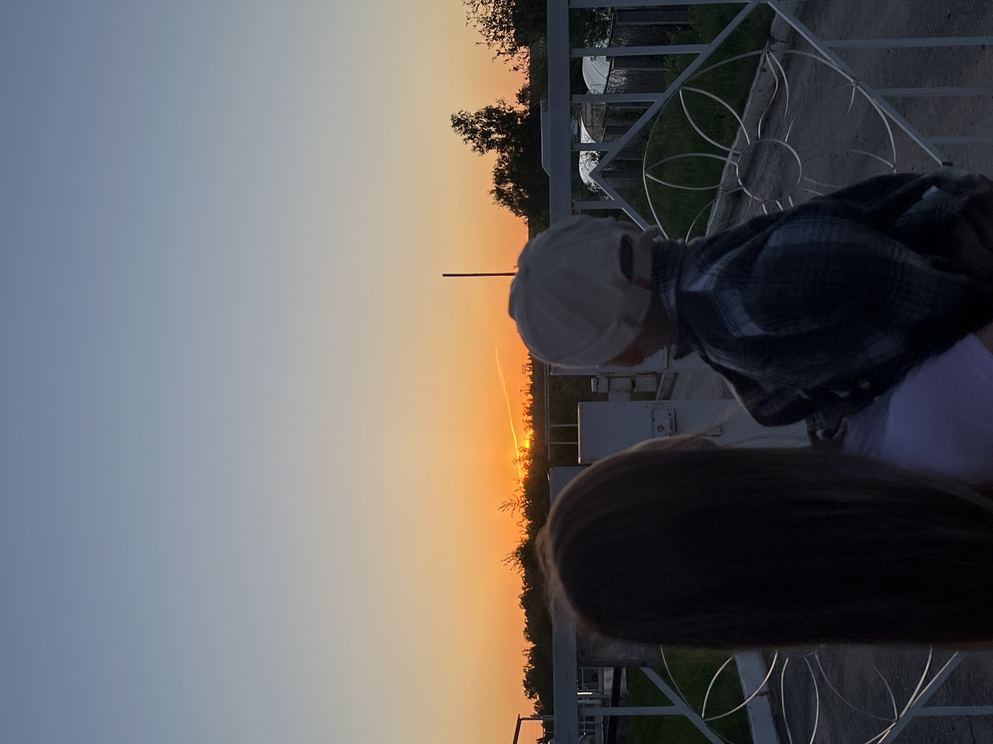 A slightly rotated outdoor shot captures the moment someone, possibly a child given the small stature, wearing a beige hat and a dark plaid shirt looks out over a setting sun. Another individual with long, dark hair stands adjacent. The scene includes a white, decorative fence and glimpses of green vegetation. The location is Фоки, Russia, indicated by metadata. In the background, further details include distant trees and some structures, potentially greenhouses. The sky displays a gradient from light blue to a warm orange hue as the sun dips towards the horizon.