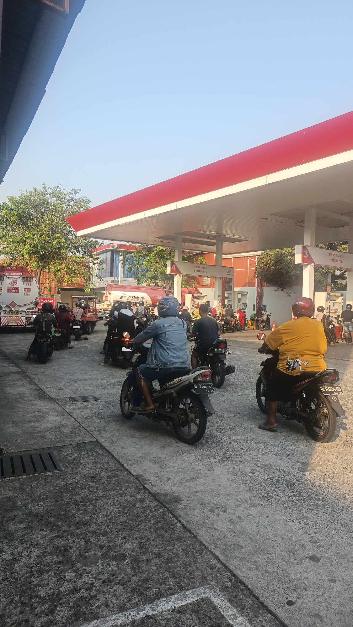 In Rendeng, Indonesia, a busy Pertamina gas station is captured from a low angle. The scene is dominated by a bright red and white canopy covering multiple fuel pumps. Several people on motorcycles are waiting in line to refuel, with some actively riding their bikes. A large Pertamina tanker truck is visible in the background, indicating the delivery of fuel. The ground is a rough concrete surface, with visible drainage grates. The sky is clear and blue, suggesting it is daytime. The overall atmosphere is one of a typical day at a busy, functional gas station.