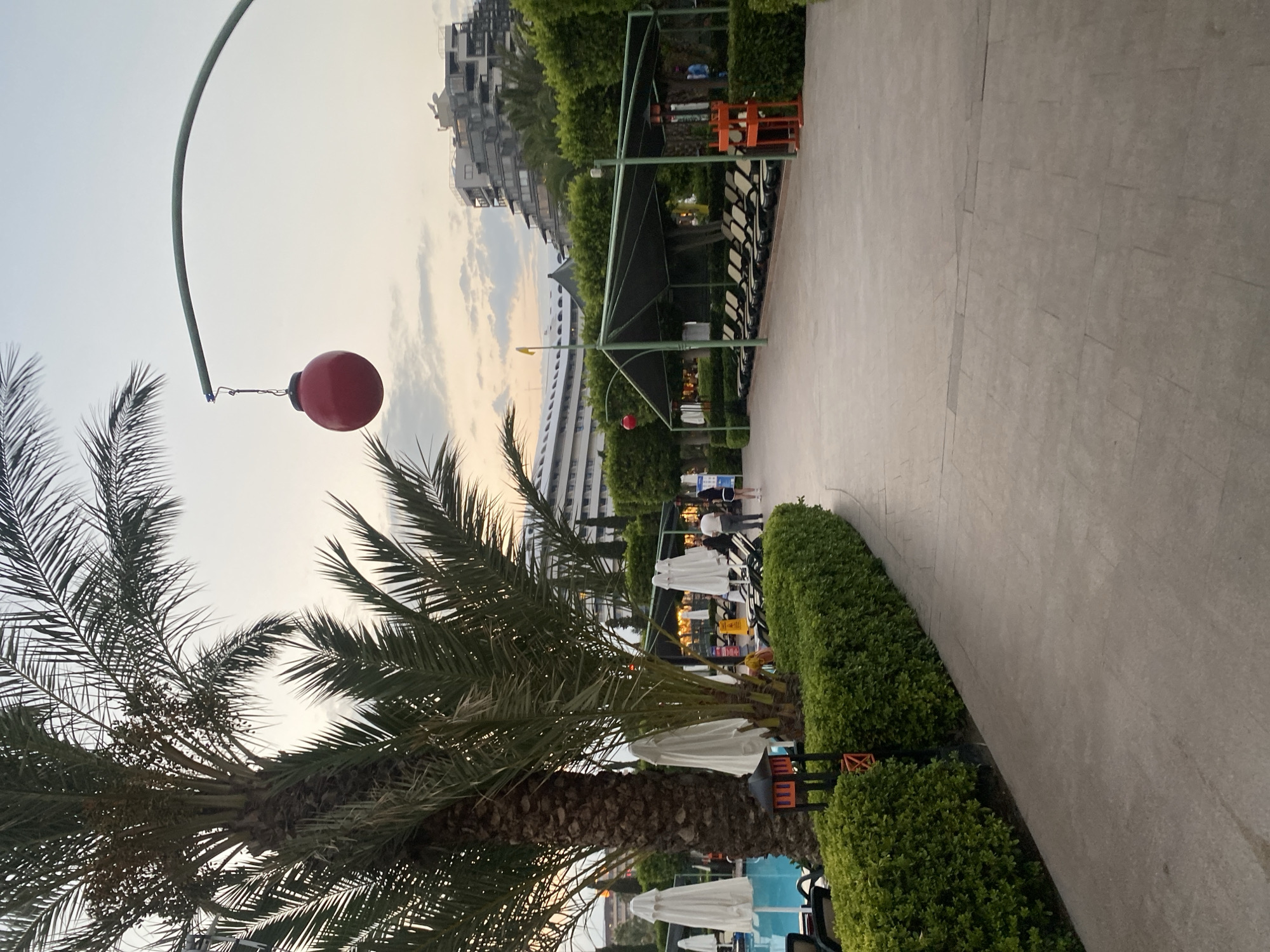 An outdoor pathway, paved with rectangular concrete slabs, curves to the right in the foreground. On the left side of the pathway, a lush green hedge runs parallel, bordering a swimming pool with blue water. Several closed white umbrellas are positioned near the pool, and a tall palm tree with visible fronds stands to the left of the path, with a small, dark red lantern-like structure at its base.
Beyond the hedge, a section of lounge chairs are visible, some covered by a dark green canopy supported by poles. A bright red, spherical ornament hangs from a curved green pole extending over the path. In the background, several multi-story buildings with modern architecture are visible, some appearing to be hotels or resorts. The sky is overcast with soft clouds, and the light suggests either early morning or late evening. Two people are seen walking in the middle ground, and another person is standing near an orange structure on the right side of the image. The scene appears to be a resort or hotel area in Aksu, Turkey.