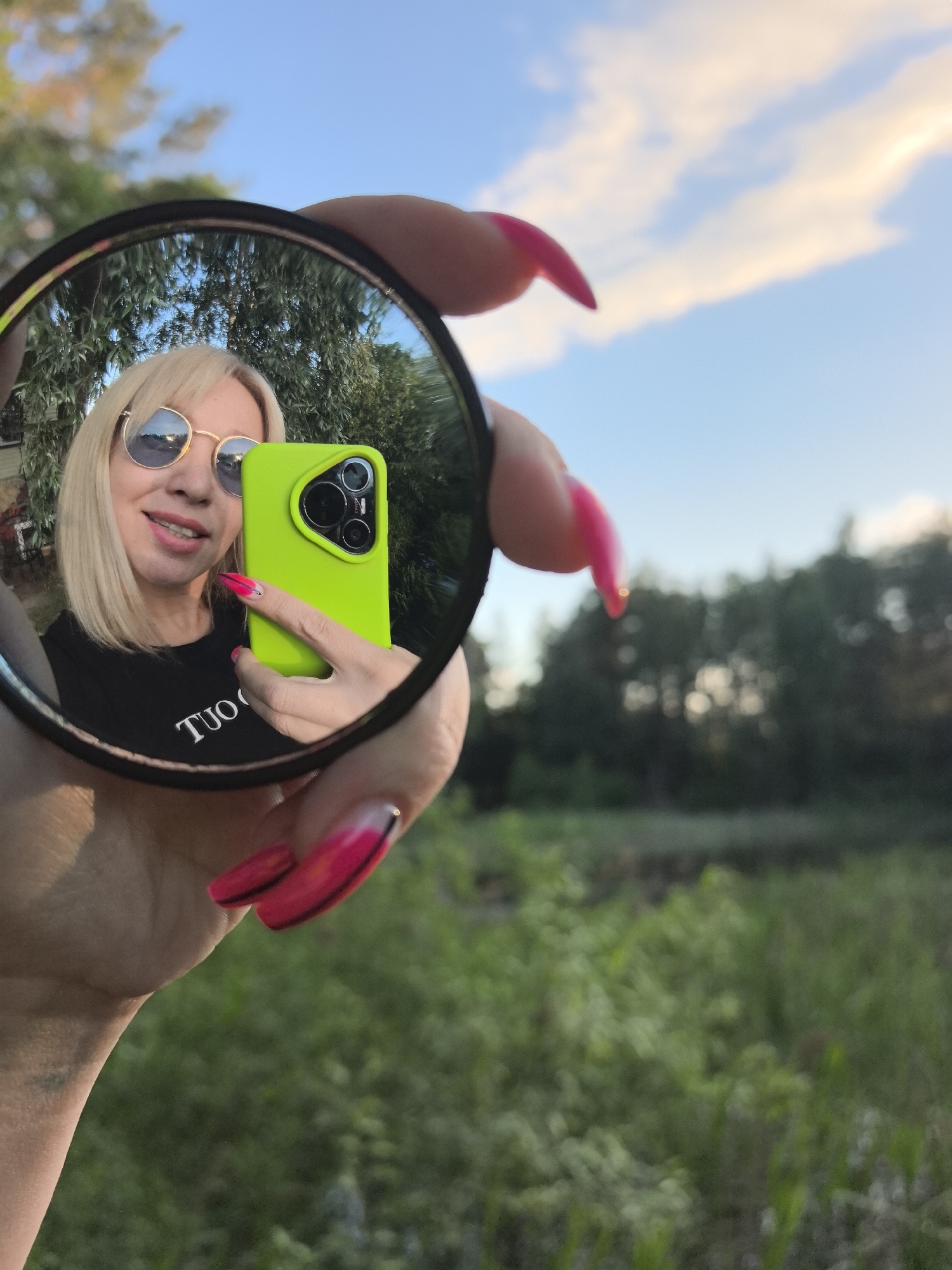 A woman with blonde hair and sunglasses takes a selfie in a mirror held in her hand. She is wearing a black t-shirt and has bright pink painted nails. In her reflection, she is holding a neon green smartphone. The background is a blur of green foliage and a blue sky with some clouds. The image was taken in the city of Noviy, Russia.