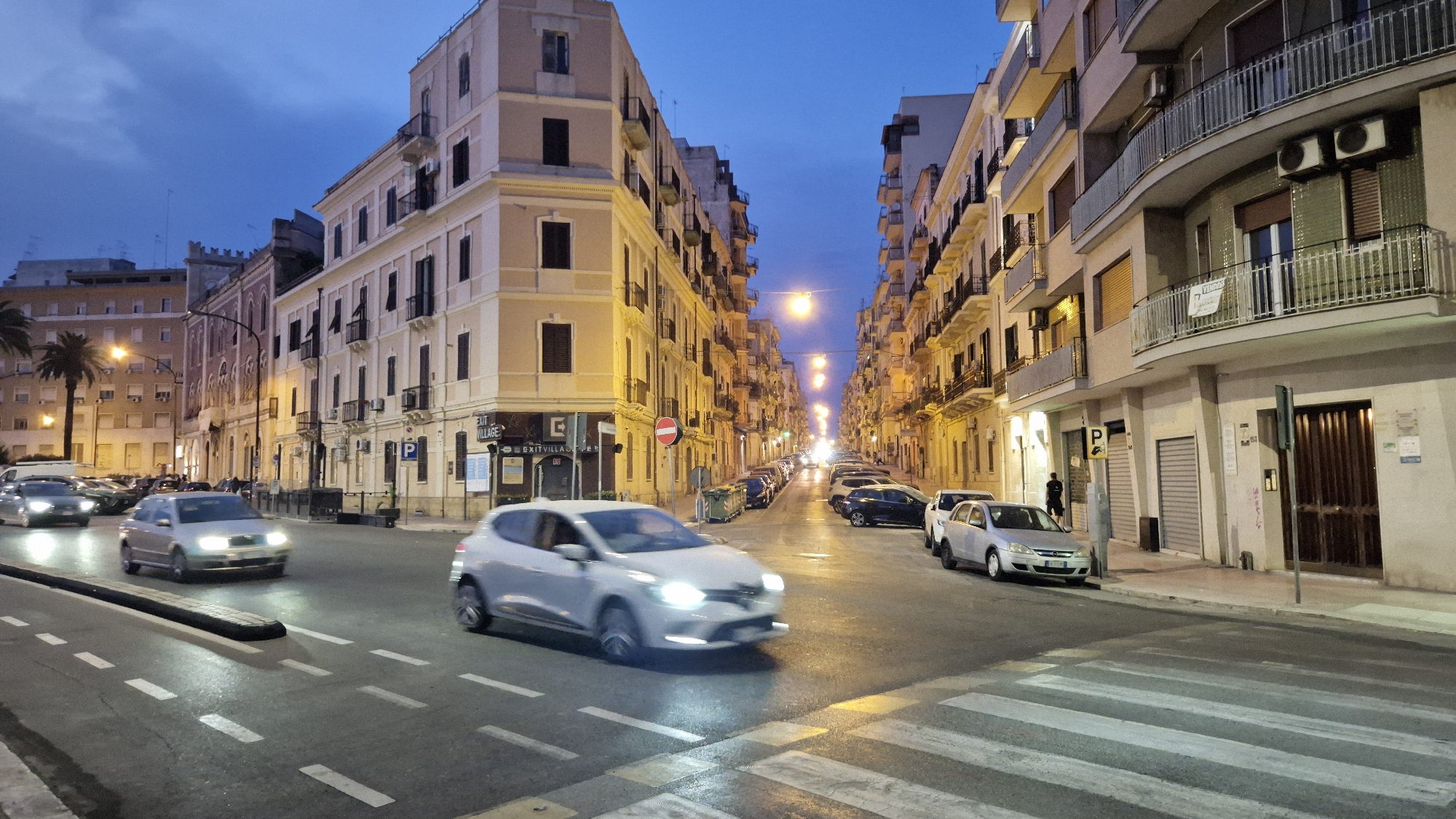 This is a wide-angle, low-angle street-level photograph of a city street in Taranto, Italy, likely taken at dusk or dawn due to the twilight sky and illuminated streetlights. The scene is dominated by buildings with neoclassical architectural styles, featuring multiple stories, balconies with railings, and shuttered windows.
In the foreground, a wet asphalt road with white lane markings and a pedestrian crossing is visible. Several cars are present on the road. A silver hatchback is in motion, crossing the pedestrian area from left to right, with its headlights illuminated. To the left, another silver car is parked, partially obscured by the moving vehicle. Further down the street, more cars are parked along the right side.
The buildings lining the street are of varying heights and colors, primarily cream and yellow, with some darker accents. Streetlights are visible at intervals, casting a warm glow on the wet pavement. Signs are present on some buildings, including a "P" for parking and a sign for "EXIT VILLAGE." A few trash bins are also visible along the side of the road. In the distance, the street extends into a vanishing point, lined with more buildings and illuminated by streetlights. A single person, appearing as a dark silhouette, is standing on the sidewalk to the right of the image, near a parked car and a street pole. Palm trees are visible in the background on the left side of the image, suggesting a Mediterranean climate. The overall impression is a typical European urban street scene during a time of transition between day and night.