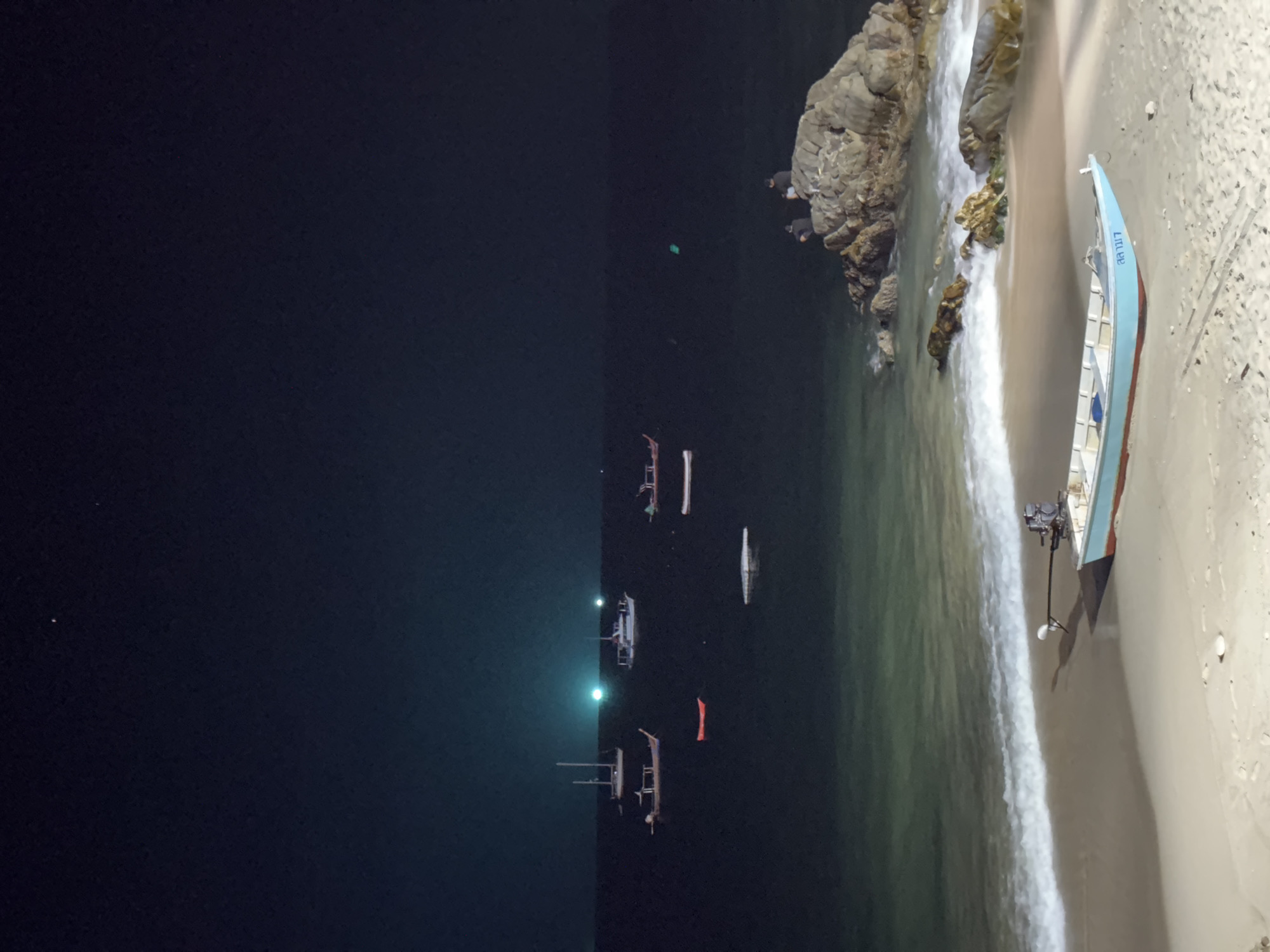 The image depicts a nighttime scene at a beach in Patong, Thailand. In the right portion of the image, there is a light blue boat resting on the sand near the water's edge, with waves lapping onto the shore. To the left of the boat, there is a rocky outcrop with people on it. Farther to the left, in the ocean, several boats are moored, their lights faintly visible. The rest of the scene is dominated by the dark expanse of the night sky.