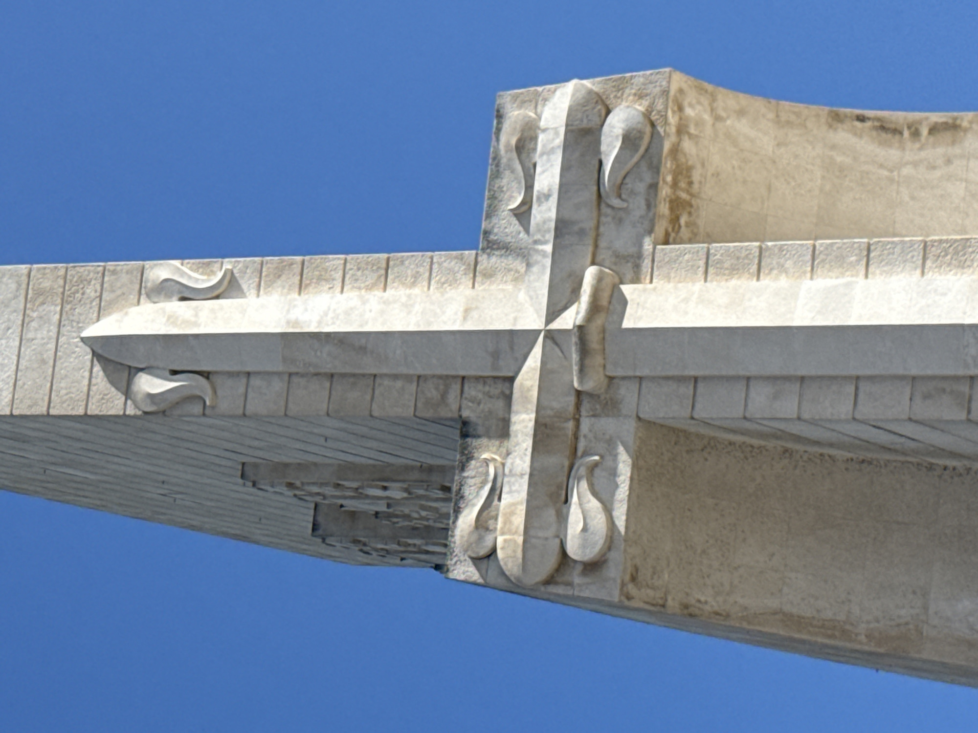 This is a close-up, low-angle, exterior, eye-level shot of a monumental concrete structure against a clear, vibrant blue sky. The structure appears to be a cross or a similar symmetrical architectural element, possibly part of a monument or a religious building. The material is a light-colored concrete, with some areas showing signs of aging and staining, particularly on the upper horizontal beam. The architectural style is modernist, characterized by clean lines and geometric forms, with decorative elements carved into the concrete.
Specifically, the image focuses on the intersection of two beams. The vertical beam features three decorative carvings, each resembling a stylized teardrop or flame shape. Two are positioned on the upper part of the beam, and one is below, all appearing to be symmetrical. The horizontal beam on the left also has decorative elements, two of which are visible, also in a teardrop or flame-like shape, facing outwards from the central intersection. The underside of the horizontal beam shows a pattern of stacked, angled blocks, creating a textured, almost tiled appearance. The surface of the concrete is textured, with visible lines from the formwork and some rough patches.
The bright blue sky indicates it is daytime, likely sunny. The clear sky suggests fair weather. The architectural style and the grandeur of the structure imply it's a significant landmark, potentially in Lisbon, Portugal, as indicated by the location context. There is no visible text in the image. No people are present. The overall impression is one of solid, enduring architecture against a bright, open sky.