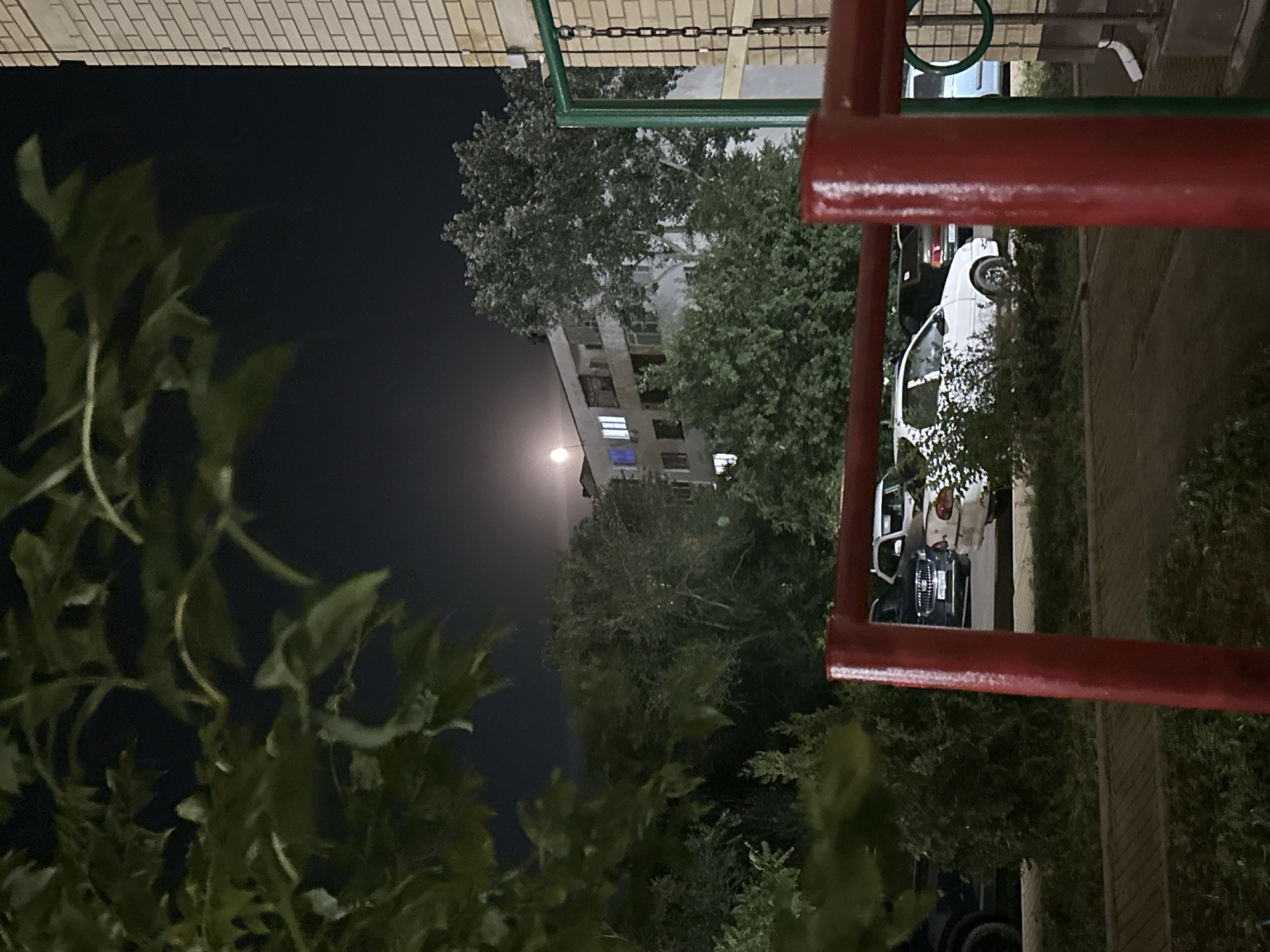 The image depicts a nighttime scene in Nevinnomyssk, Russia. In the foreground, a red metal railing is visible, with a green metal chain link fence to the right. Beyond the railing, several cars are parked. A white sedan is prominent in the center, with a dark-colored SUV to its left. Further back, a multi-story apartment building with illuminated windows is visible, partially obscured by trees. The moon is faintly visible in the night sky above the building. The overall environment suggests a residential area at night.