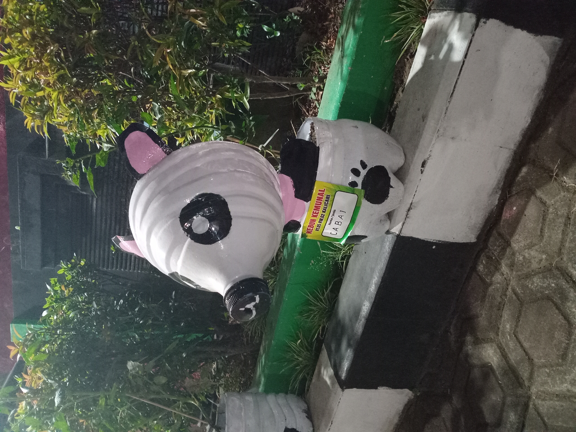 A white, panda-like figure made from recycled plastic bottles sits on a concrete planter box in what appears to be an urban garden setting at night. The figure has black eyes, a black snout, and pink ears. A yellow label is attached to the front of the figure, with text including "KEBUN KEMUNAL" and "RT.05 RW.02 KALICARI", indicating it is located in the Kalicari neighborhood (RT 05, RW 02) in the city of Semarang, Indonesia. The planter box is painted with black and white stripes. Behind the planter box, lush green foliage is visible, along with a dark, corrugated wall. The ground in front of the planter box is paved with interlocking hexagonal concrete stones. The lighting suggests it is dark, possibly early morning or evening.