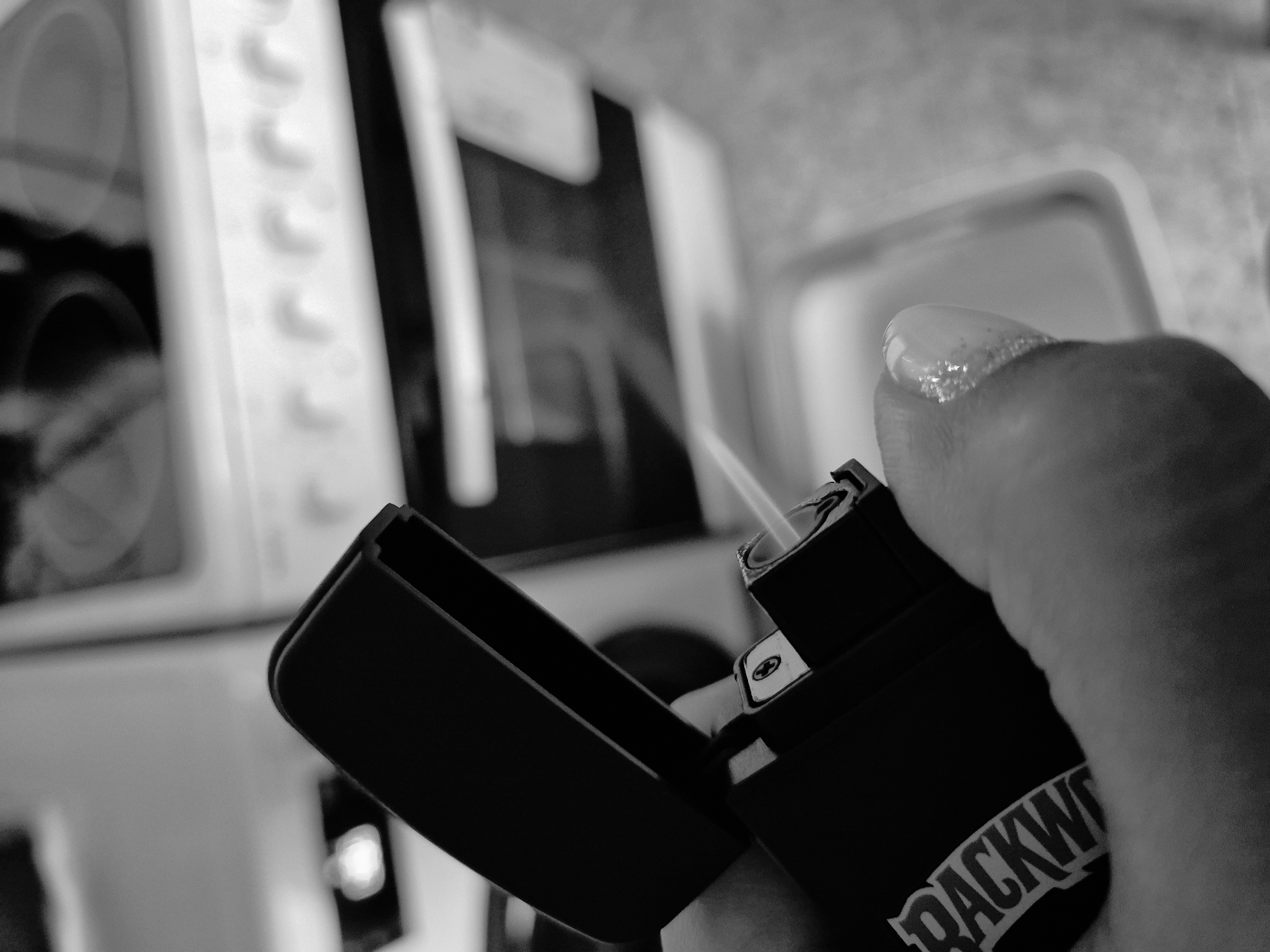 This monochromatic image captures a close-up, high-angle view of a human hand holding an activated, open, black rectangular device, resembling either a Zippo-style lighter or a pod vape. The hand, specifically a thumb and index finger, is visible in the foreground, with the thumb's fingernail displaying a clear or light-colored, textured, possibly glittery, acrylic coating. The device has a hinged lid that is open upwards, revealing internal components, including a Phillips head screw on a metallic element. A thin, white stream of light or vapor is emitted from an aperture within the device, indicating its active state. Partial white lettering, "BACKW," likely part of "BACKWOODS," is visible on the side of the device.
The background, rendered out of focus, reveals a kitchen environment. A white kitchen range or stove is prominently visible, featuring a row of circular control knobs on its front panel, and a glass cooktop with blurred circular burner outlines above. An oven door is discernible below the controls. To the lower right, another indistinct kitchen surface, possibly a countertop or sink, is present. The scene is illuminated by ambient indoor light. The reported location of this scene is Iskrets, Bulgaria.