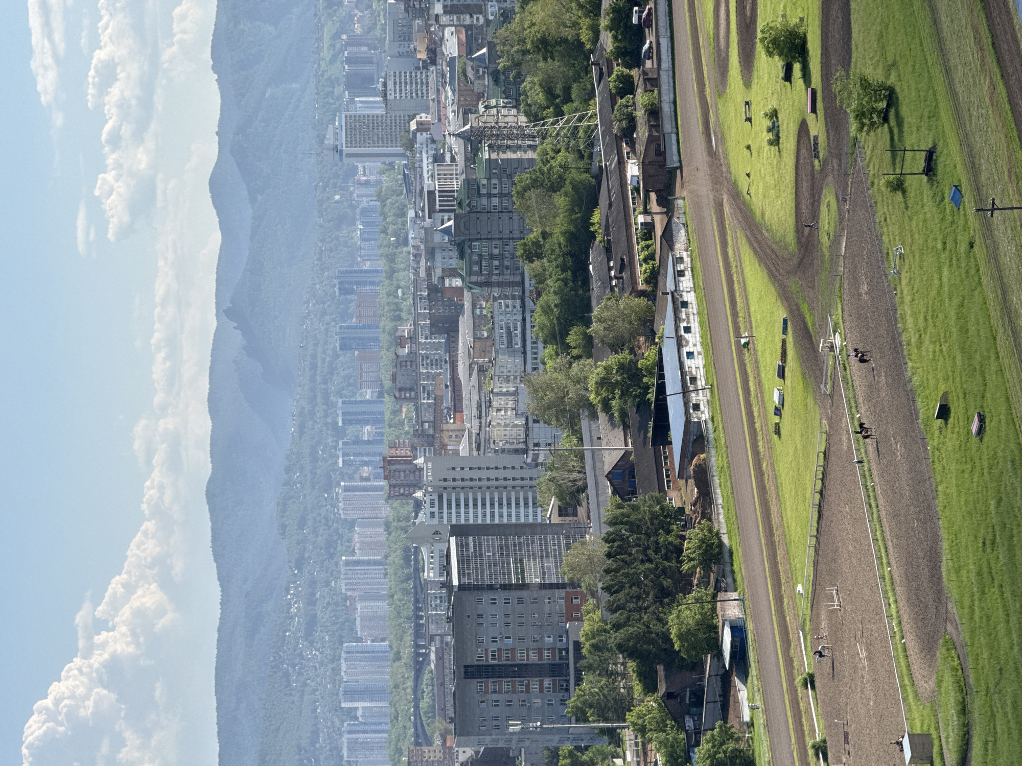 The image shows an aerial view of a cityscape with mountains in the background. In the foreground, there is a large equestrian facility with several fenced-off dirt arenas. Some horses and people are visible within the arenas, with some individuals riding horses. Surrounding the arenas are patches of green grass and dirt paths. In the middle ground, there are various buildings, including what appear to be industrial or agricultural structures, as well as some residential buildings. Beyond these, a dense collection of high-rise apartment buildings stretches across the urban landscape. The distant background is dominated by a range of hazy, blue mountains under a bright sky with scattered white clouds. The lighting suggests it is daytime.