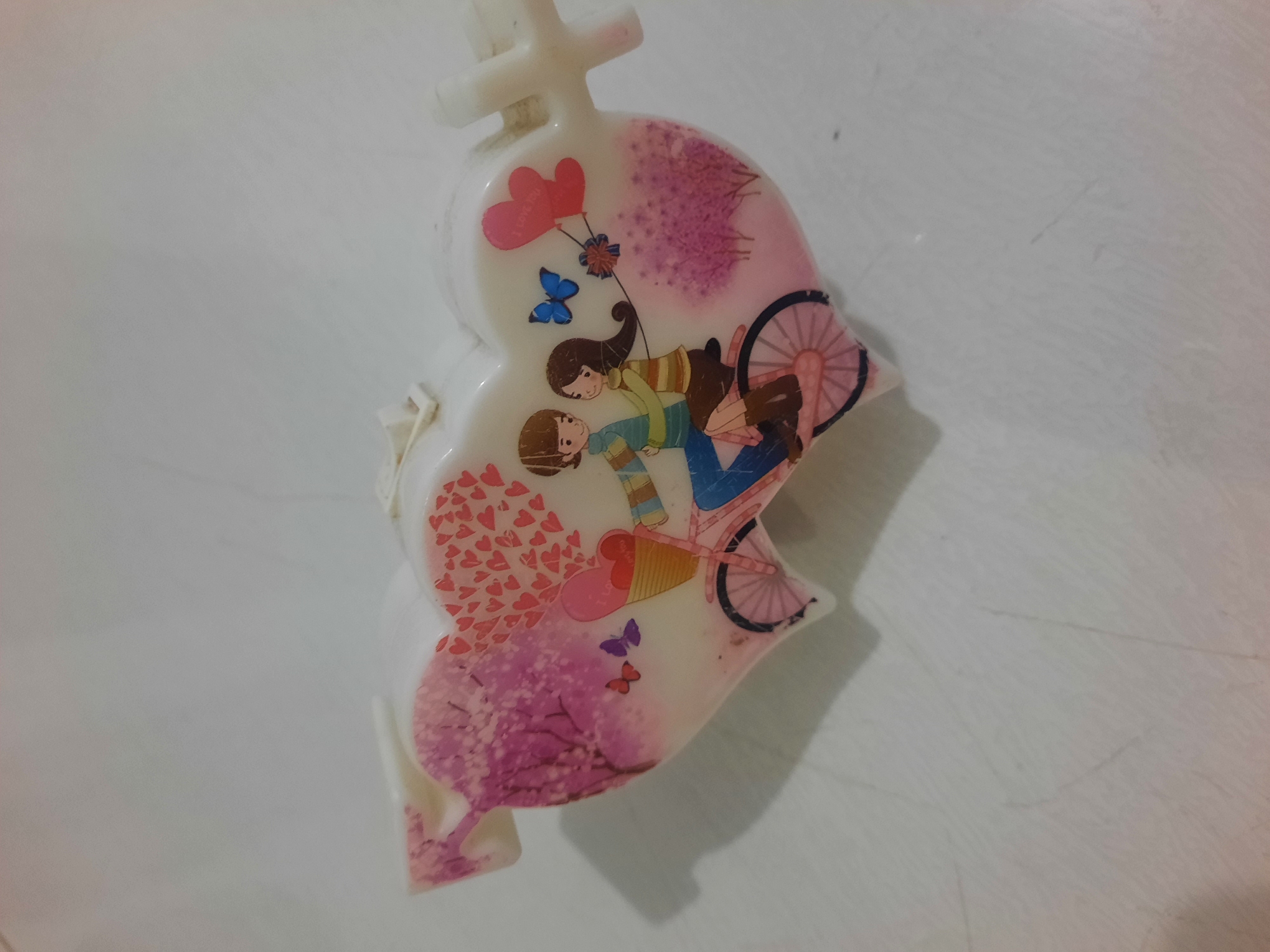 The image shows a heart-shaped object with a printed illustration of a couple riding a bicycle. The illustration features a man and a woman, both with dark hair, on a pink bicycle. The man is wearing a blue and green striped shirt, and the woman is wearing a brown and yellow striped shirt and a brown skirt. They are surrounded by decorative elements such as pink trees, butterflies, and balloons. The heart object is white with pink and red accents. The object appears to be a candle or a decorative trinket. The background is a light-colored, textured surface, possibly a table or countertop, with some subtle shadows. There is no indication of the city of Lhokseumawe, Indonesia in the image.