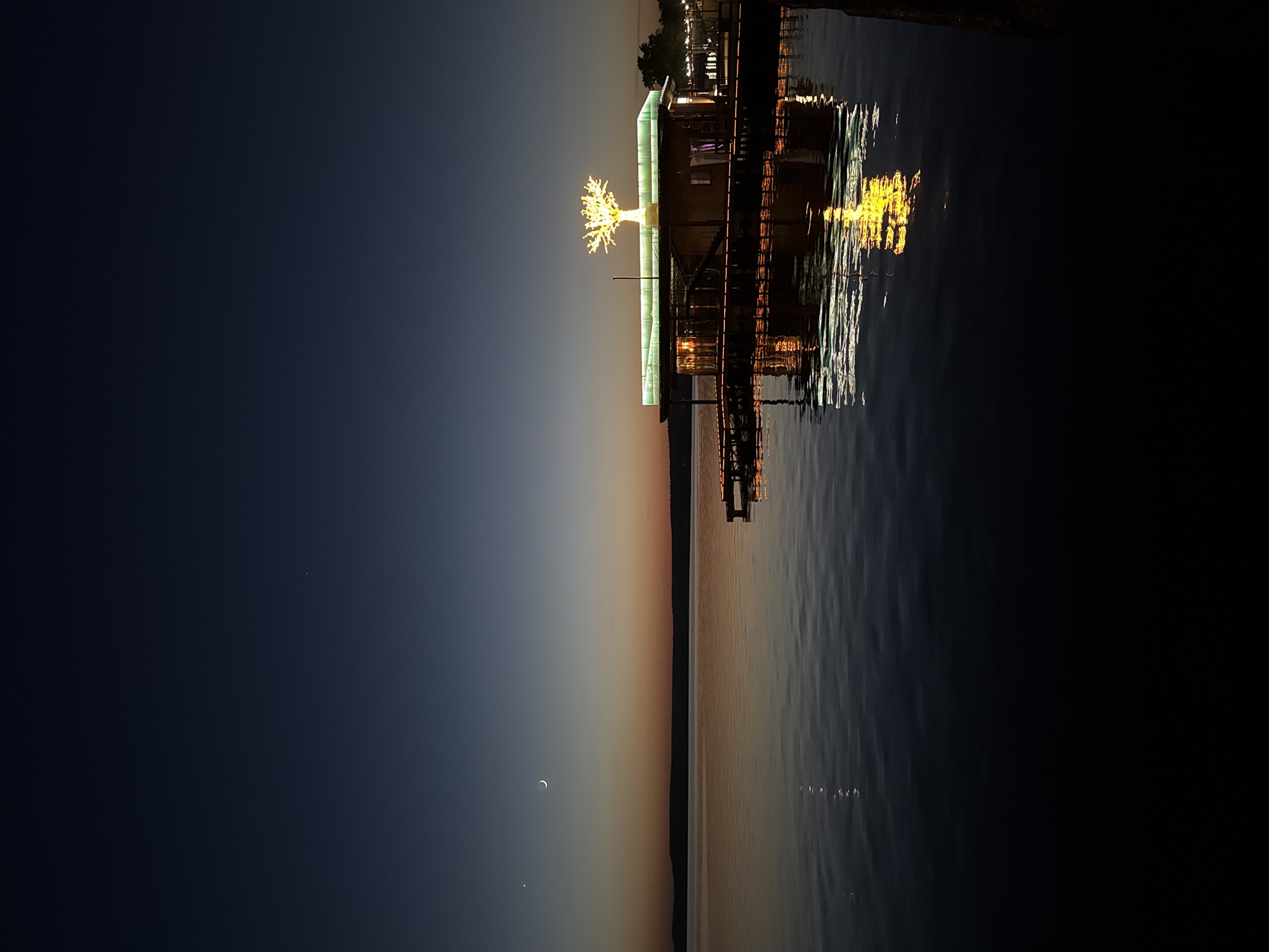 The image shows a waterfront scene at dusk in Votkinsk, Russia. A structure, possibly a pier or a building extending over the water, is illuminated with lights, including a decorative tree-like fixture. The lights are reflected in the water's surface, creating shimmering patterns. In the sky, a crescent moon and a single star are visible above the horizon. The sky transitions from a dark blue to a muted orange near the horizon. The overall scene depicts a peaceful evening atmosphere on the waterfront.