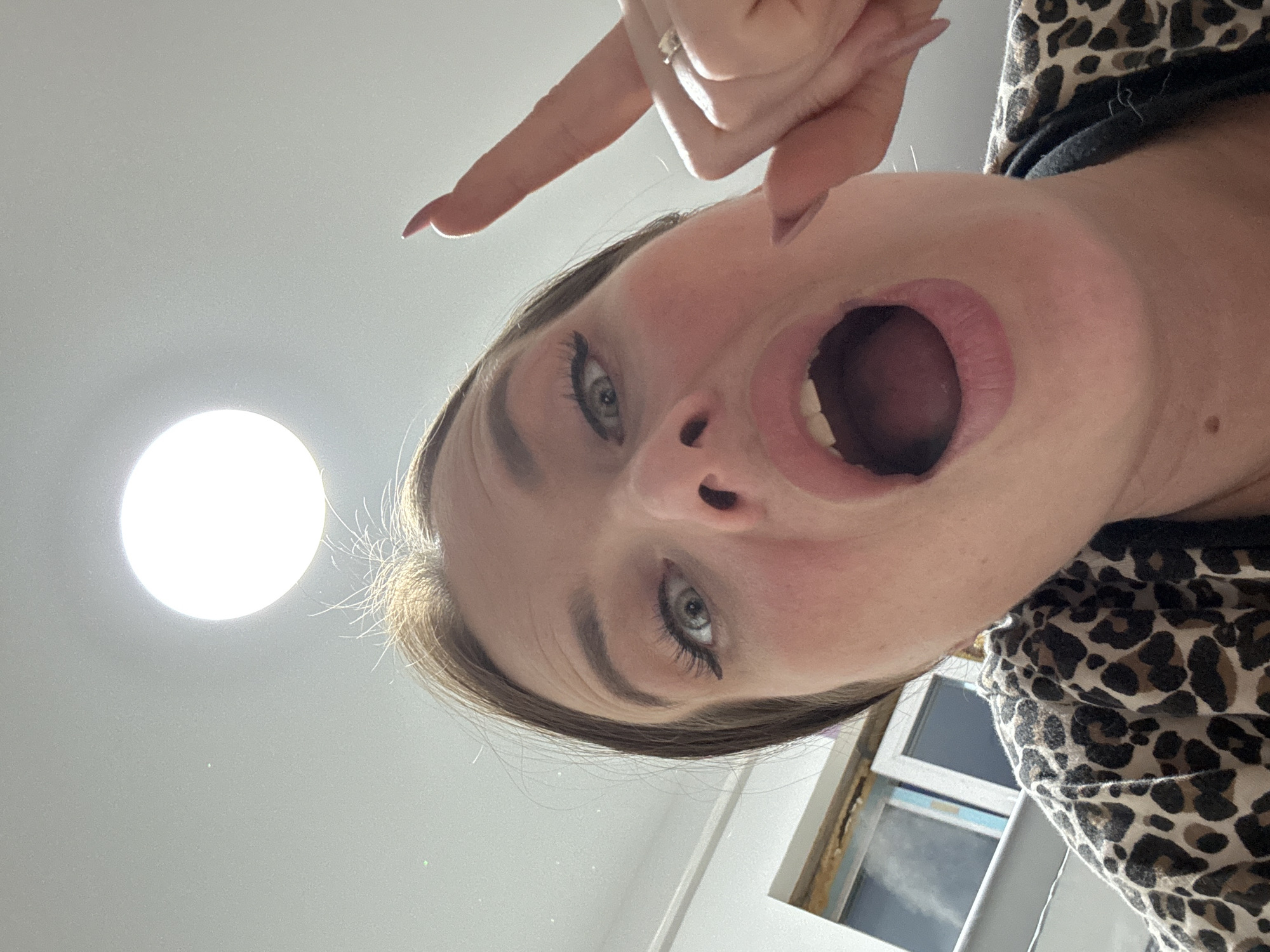 A woman is holding her finger and pointing to her open mouth with a light in the background, wearing a leopard-print shirt. A window is visible in the background. The location of Rusalevka, Russia, cannot be determined from the image.