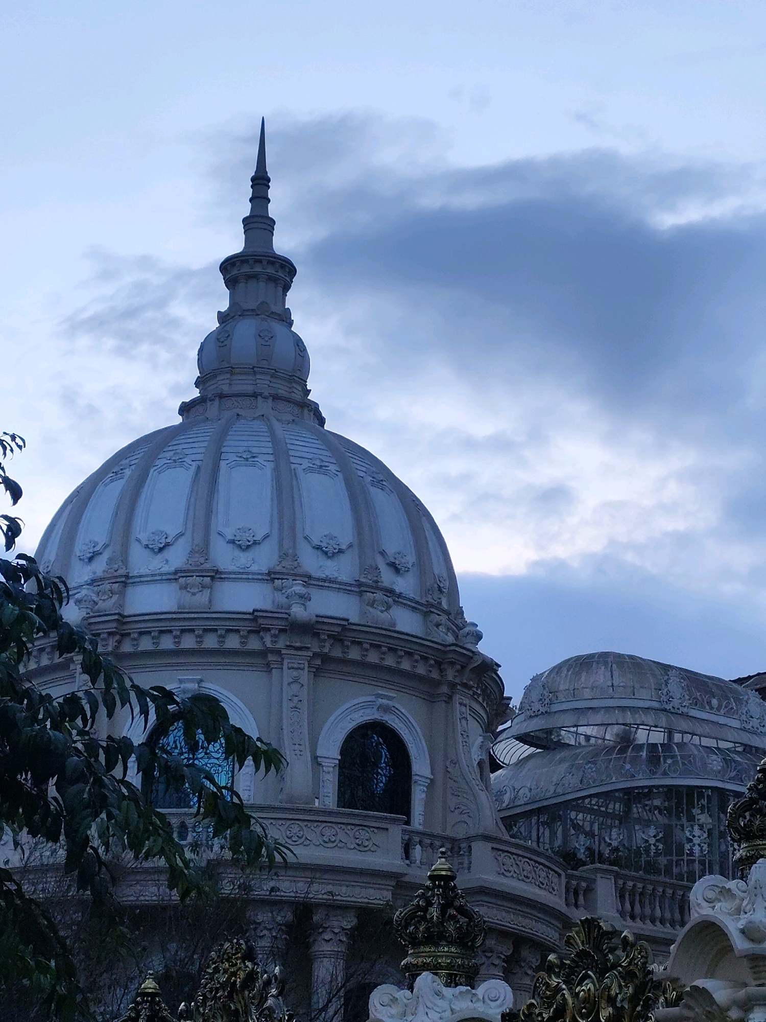 The image shows a close-up view of a domed building with ornate architectural details, possibly a part of the Bao Dai Summer Palace in Da Lat, Vietnam. The building features a large, pale dome topped with a spire, surrounded by decorative moldings and sculptural elements. In the background, another glass-domed structure is visible, suggesting a conservatory or greenhouse. Dark green foliage from a tree partially obscures the left side of the building. The sky is overcast with dramatic, dark clouds, creating a somber mood. The foreground is partially obscured by decorative golden and white architectural features, possibly a fence or part of the palace's exterior. The overall impression is of a grand, historic structure set against a moody sky.