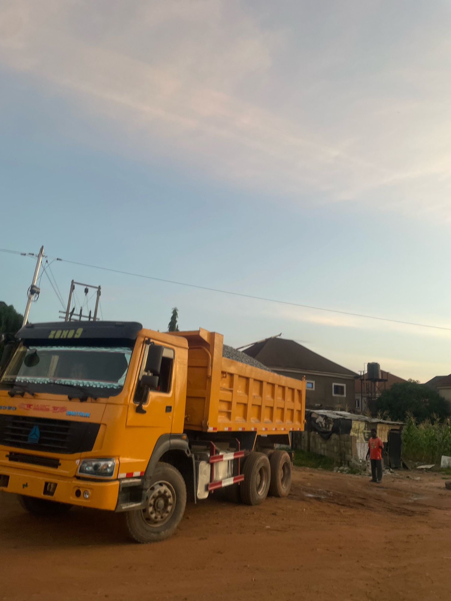 A yellow dump truck is parked on a dirt road in Abuja, Nigeria. The truck is loaded with what appears to be gravel. In the background, there are several houses, a water tank, and some vegetation. A man in an orange shirt is walking away from the truck. The sky is clear with a few wispy clouds. The lighting suggests it is either early morning or late afternoon. The overall scene depicts a construction or hauling site in a residential area of Abuja.