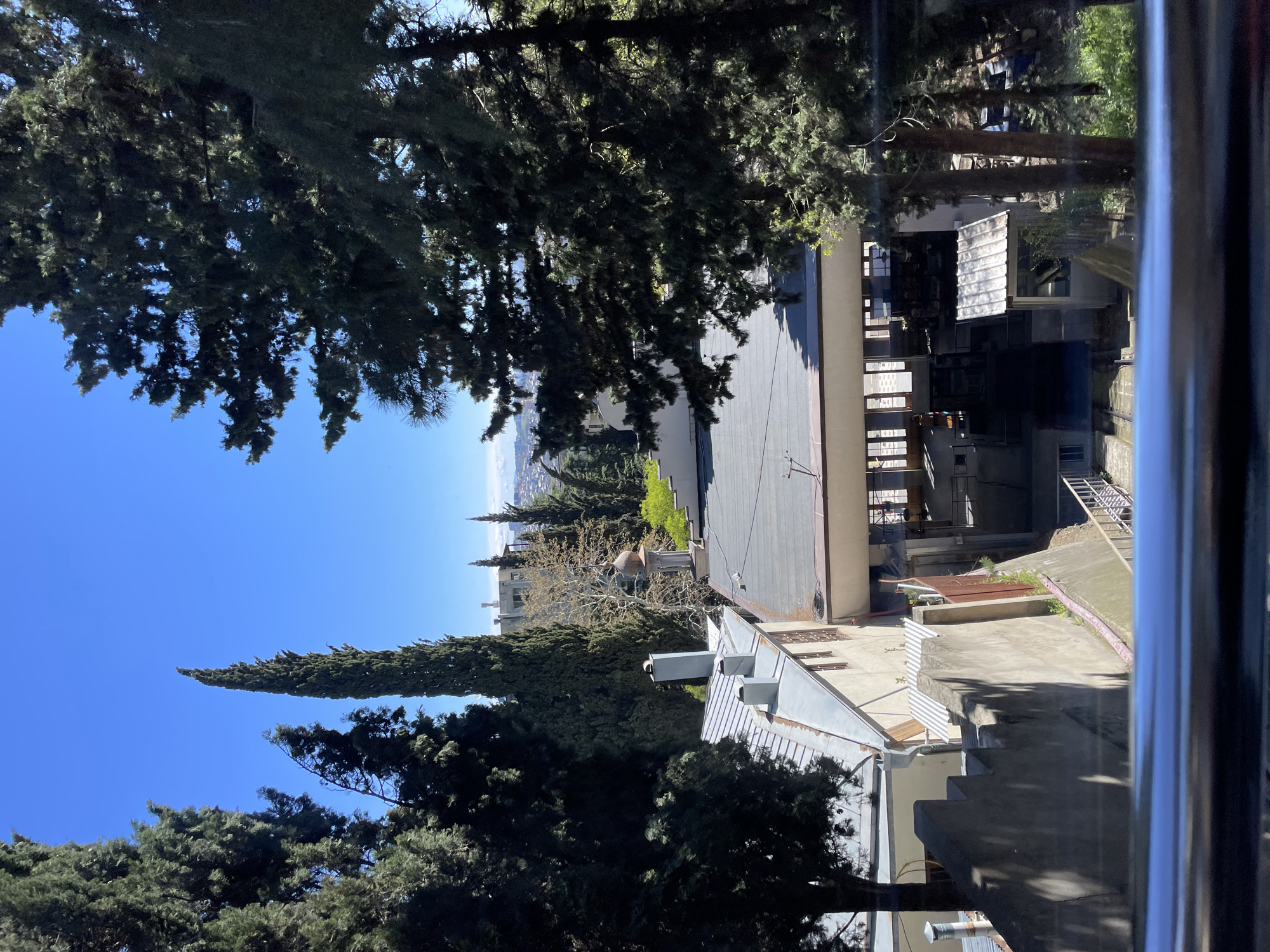 The image shows a view from a window, likely in Tbilisi, Georgia, looking out over a building complex and towards a cityscape in the distance. A blue sky is visible above the buildings and trees. The architecture seems to be a mix of older and more modern styles. Trees are abundant, partially obscuring the buildings and skyline. The scene is taken during daylight, and the light suggests a sunny day.