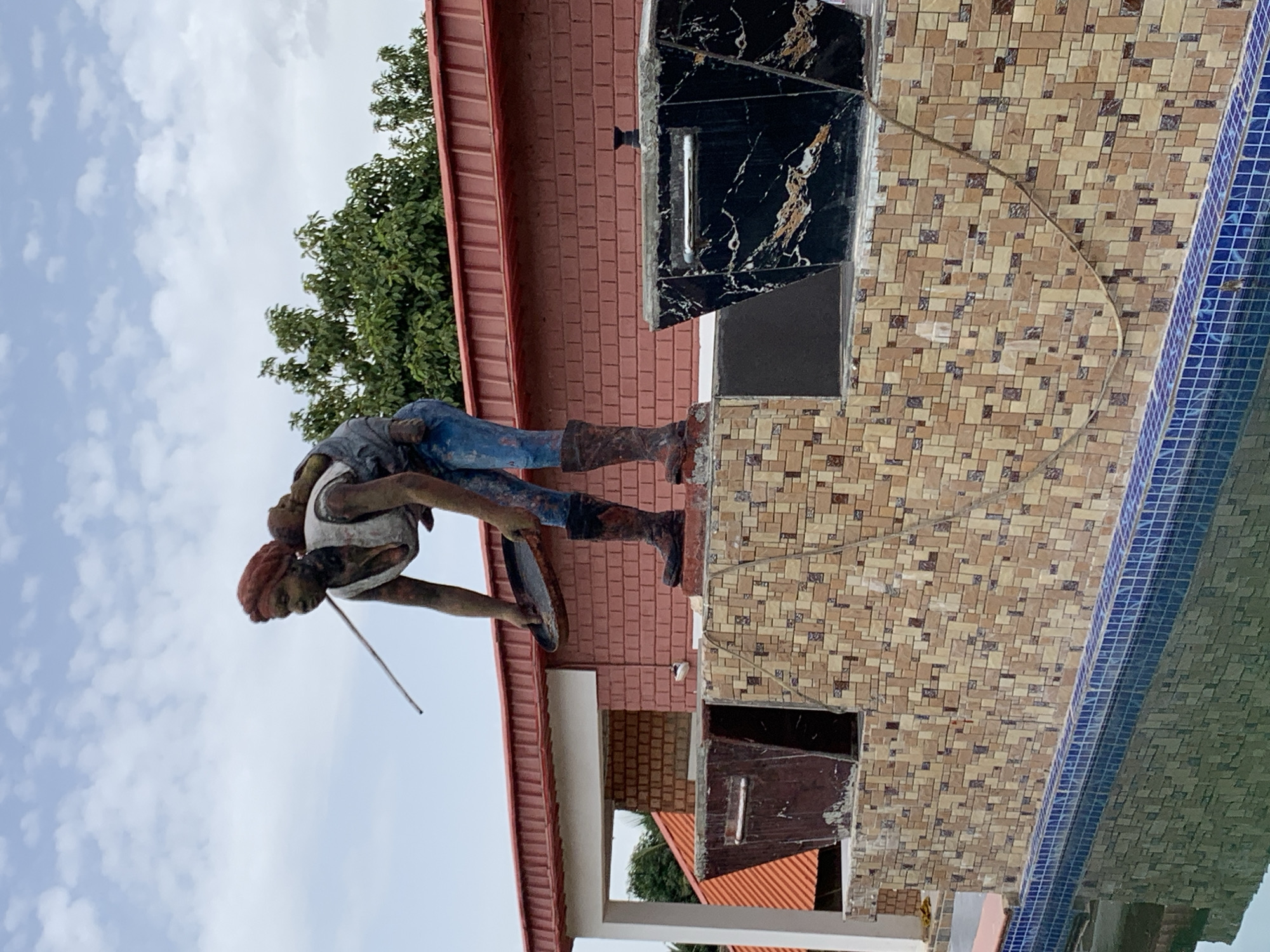 A statue of a woman carrying a child on her back, bent over a pan, is situated on a multi-level structure adorned with mosaic tiles, in front of a red-tiled roof building. The structure also features two large planters, one dark and textured, the other black with marble-like patterns. A swimming pool with blue tiled edges is visible in the foreground, reflecting the sky and the surrounding structures. The location is identified as Jos, Nigeria. The sky is partly cloudy.