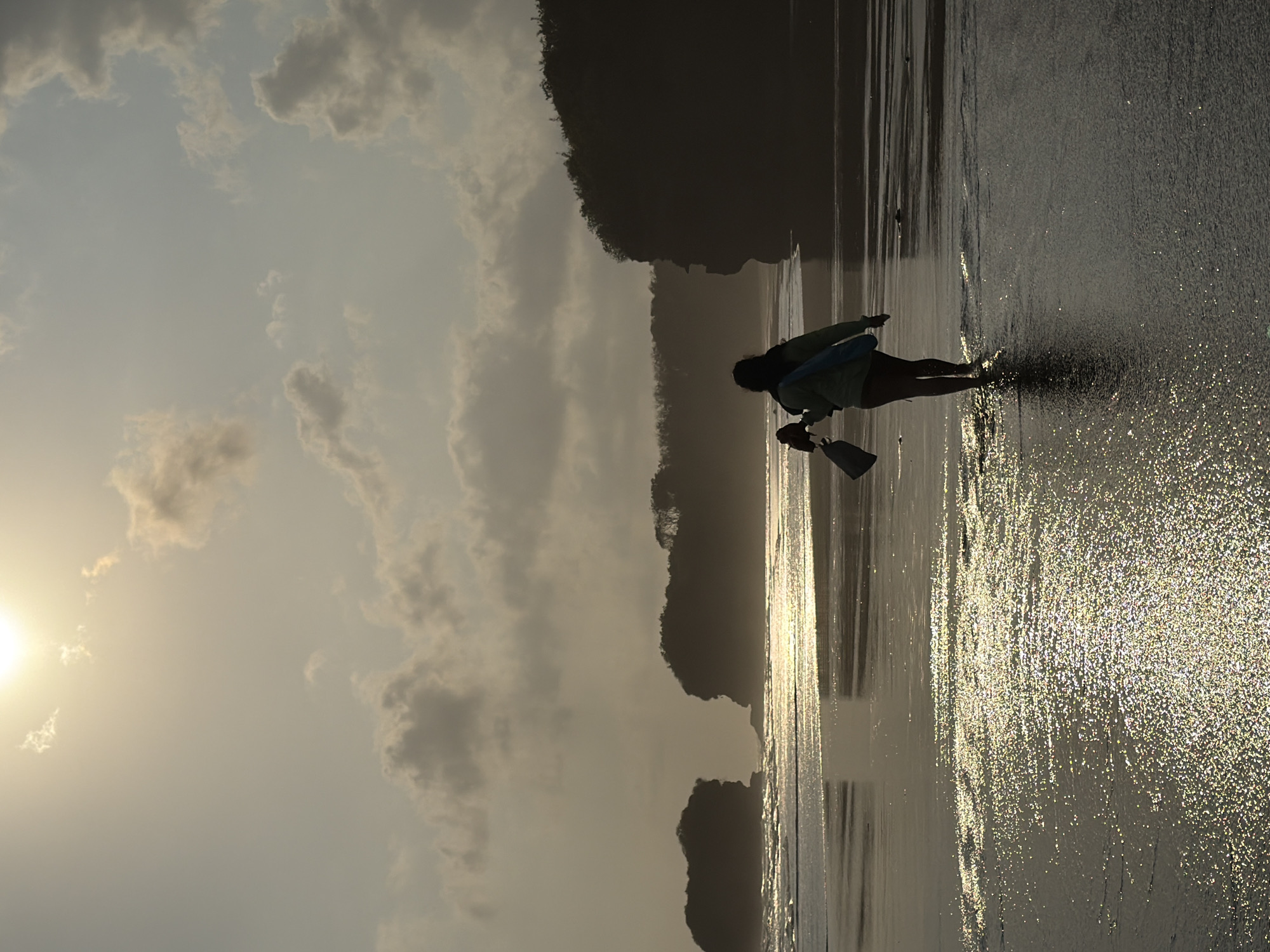 The image depicts a solitary figure walking along the shoreline at sunset in Selemadeg, Indonesia. The person, silhouetted against the bright sky, is carrying two items: one appears to be a rolled-up mat or towel, and the other a dark bag. The sun is partially obscured by clouds, casting a warm, hazy glow over the scene. The wet sand reflects the light, creating a shimmering effect. In the background, dark, tree-covered cliffs rise from the sea. The waves are gently lapping at the shore.