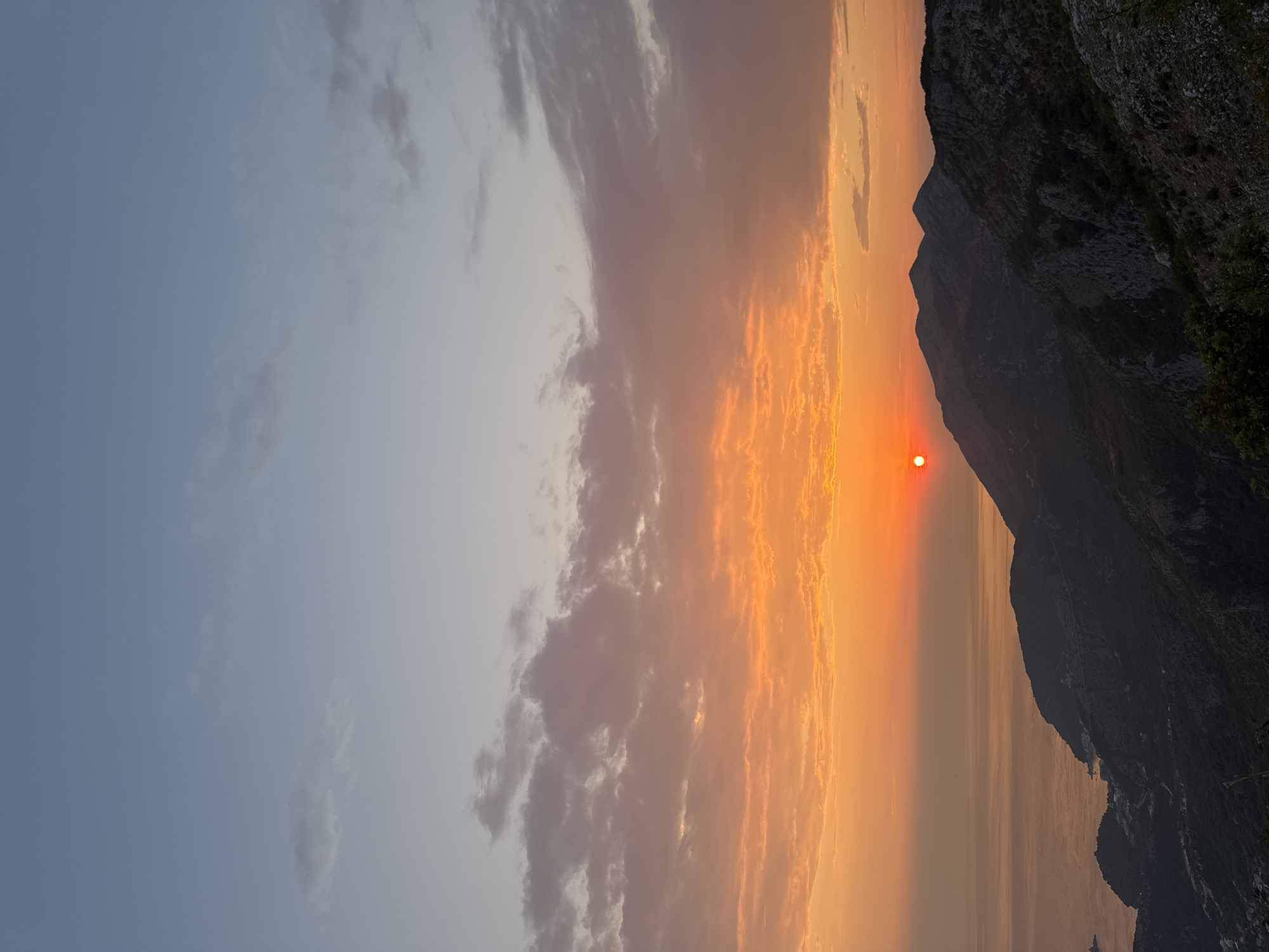 This is a vertical, full-frame photograph taken at dusk from a high vantage point overlooking the coast near Gluhi Do, Montenegro. The sky transitions from a pale blue at the top to a vibrant orange and pink hue near the horizon, illuminated by the setting sun. The sun itself is a bright orange orb positioned on the left side of the frame, near the horizon line. Scattered clouds, tinged with orange and grey, are visible in the lower half of the sky.
Below the sky, a dark, mountainous coastline dominates the lower portion of the image. The mountains are rendered in silhouette and deep shadow, with rugged terrain and sparse vegetation visible in some areas. A winding road or path is subtly discernible on one of the slopes. In the distance, the calm sea stretches towards the horizon, reflecting the colors of the sunset. Small, indistinct settlements or structures are clustered along the coastline at the base of the mountains. The foreground shows the rocky, textured surface of the elevated viewpoint with some dark green foliage. There are no people visible in the image.