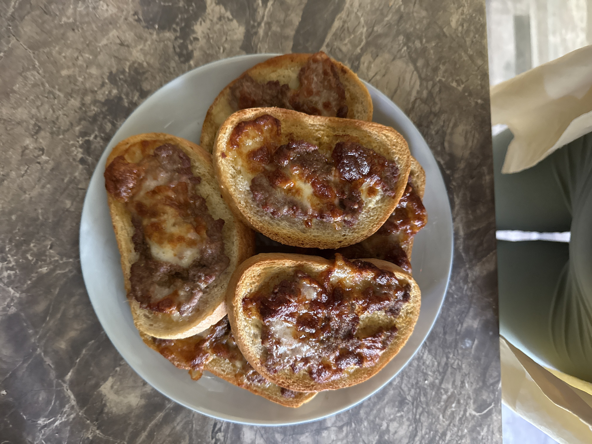A light blue plate holds several pieces of toast topped with a meat mixture and melted cheese. The toast pieces are arranged in a circular pattern, overlapping each other. The plate sits on a marble-patterned surface. The bottom portion of the image shows a person's legs and a paper bag. The scene is indoors. This image was not taken in Plovdiv, Bulgaria.