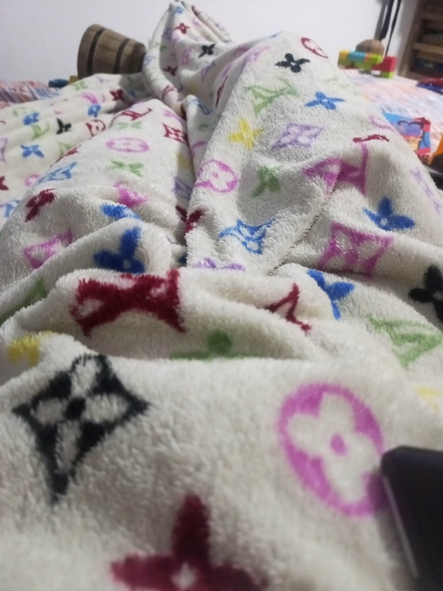 The image shows a close-up of a fluffy white blanket with a repeating pattern of colorful flowers and geometric shapes. The blanket is draped over something, creating folds and shadows. In the background, a wooden barrel is visible, along with some colorful blocks and part of a shelf. The lighting appears to be indoor and soft. The scene is a close-up perspective of the blanket, with the focus on its texture and pattern. The location in La Tagua, Venezuela, is not discernible from the visual information provided in the image.