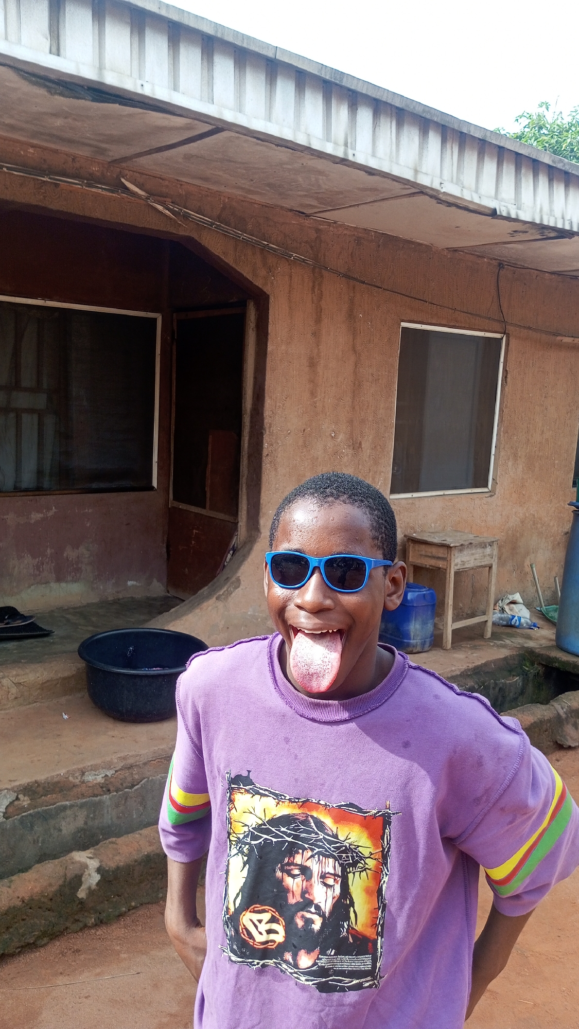 A young person with dark skin, close-cropped hair, and blue sunglasses stands outdoors, sticking out their tongue. They are wearing a light purple sweatshirt with a graphic of a man with long hair and a crown of thorns on the front. The shirt has stripes of yellow, green, and red on the sleeves. Behind the individual is a single-story building with textured, reddish-brown walls and a light-colored metal roof. The building has two windows and an open doorway. To the left of the individual is a large black basin on a concrete step. To the right, there is a small wooden table and a blue plastic container. The ground appears to be reddish-brown dirt. The location is Nkpor, Nigeria.