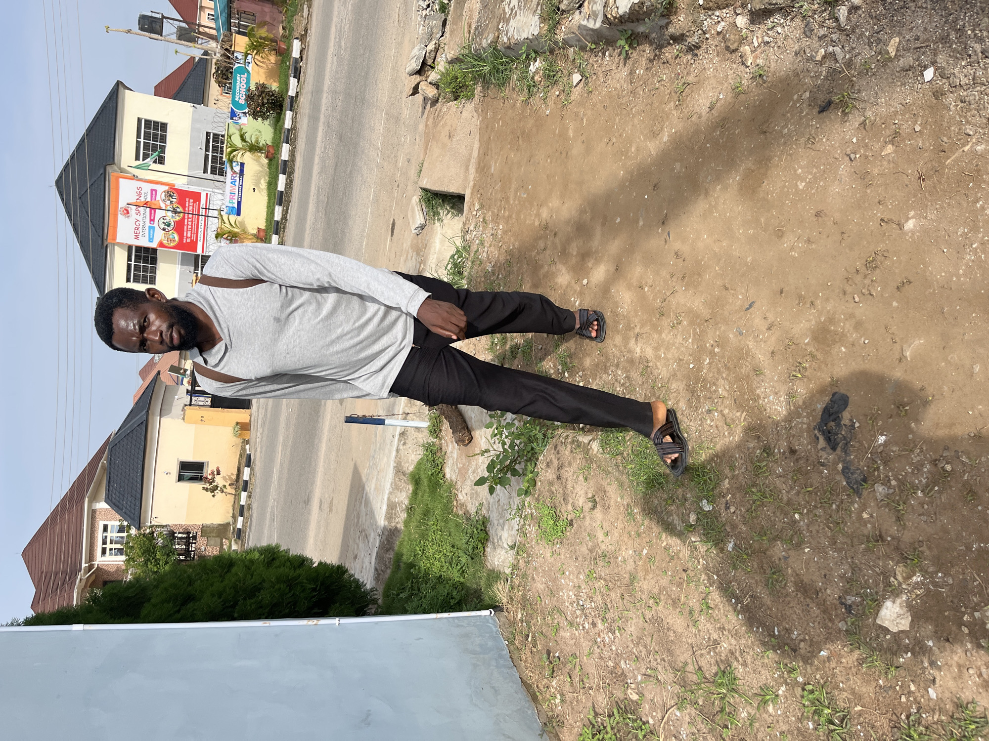 A man is standing on a dirt and grass patch beside a paved road and sidewalk. He is wearing a light grey long-sleeved shirt, black pants, and black sandals. Behind him, there are buildings, including one with a sign that says "Mercy Springs International School." The location appears to be an urban or suburban area, possibly in Abuja, Nigeria.