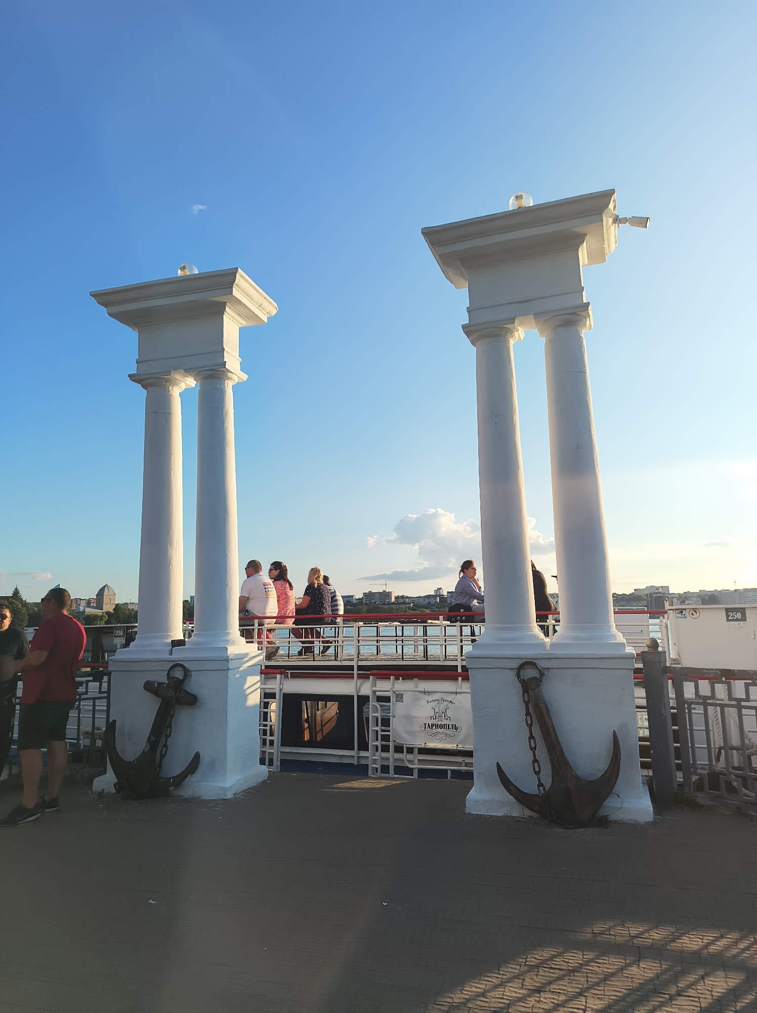 In Ternopil, Ukraine, a scene unfolds on a waterfront promenade featuring two grand white pillars with classical architectural elements, each topped with a glass orb and a small light fixture. Two large, dark anchors are prominently displayed at the base of each pillar, attached by chains. A group of people, casually dressed, are gathered on a walkway behind the pillars, suggesting a public gathering or a tourist attraction. The sky above is a clear blue, with a hint of a cloud. The overall impression is a sunny day in a scenic urban environment, likely a park or embankment by a body of water, with the architectural features serving as a landmark.