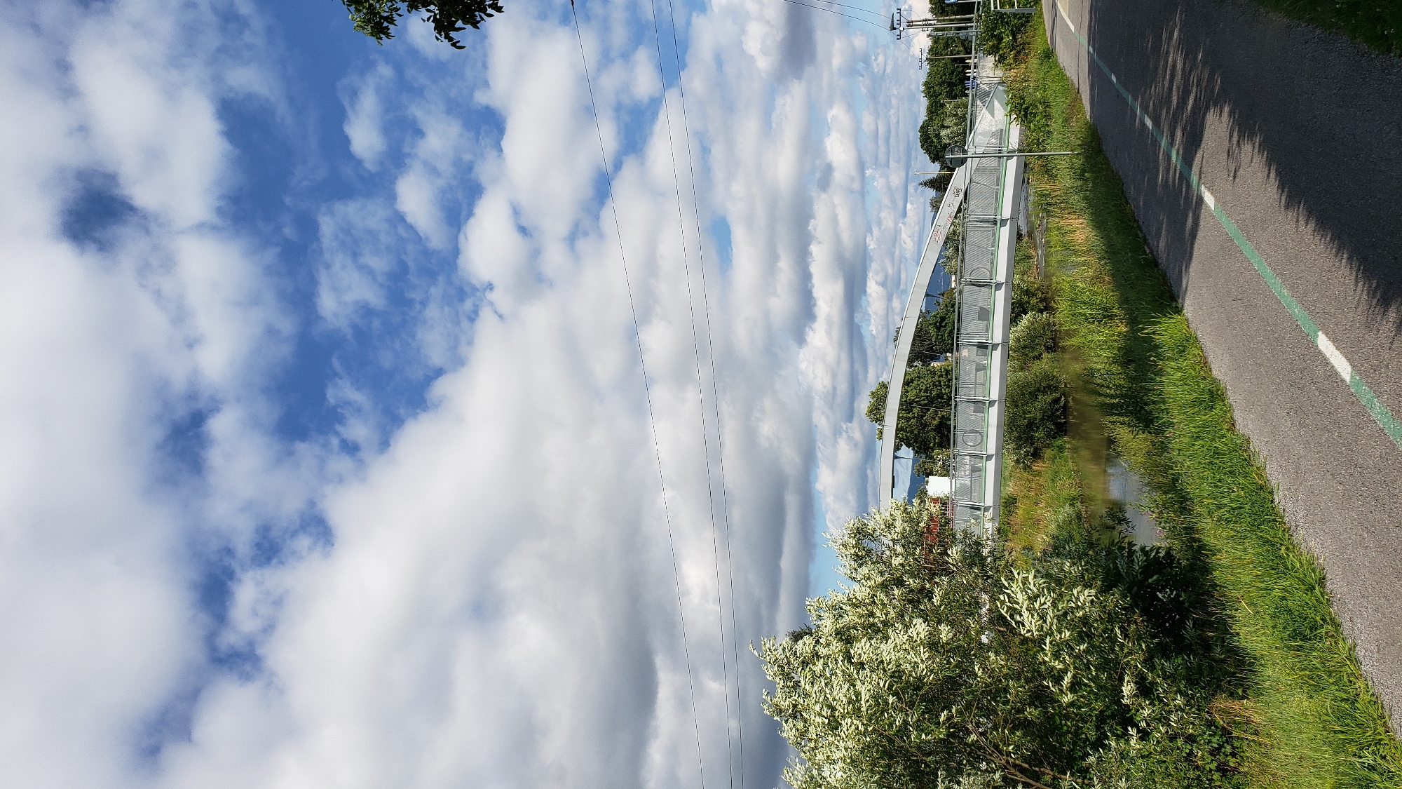 The image depicts a pedestrian and cyclist bridge spanning a river in the city of Prešov, Slovakia. The bridge, constructed with a modern, curved design, has a light-colored railing and a pedestrian walkway. It is situated amidst lush green vegetation, with trees and bushes lining the riverbanks. A paved path, likely a bike lane, is visible in the foreground, marked with a green line and white dashes. The sky is a mix of blue and white clouds, suggesting a partly cloudy day. The scene is serene and captures a moment of outdoor activity in an urban environment.
