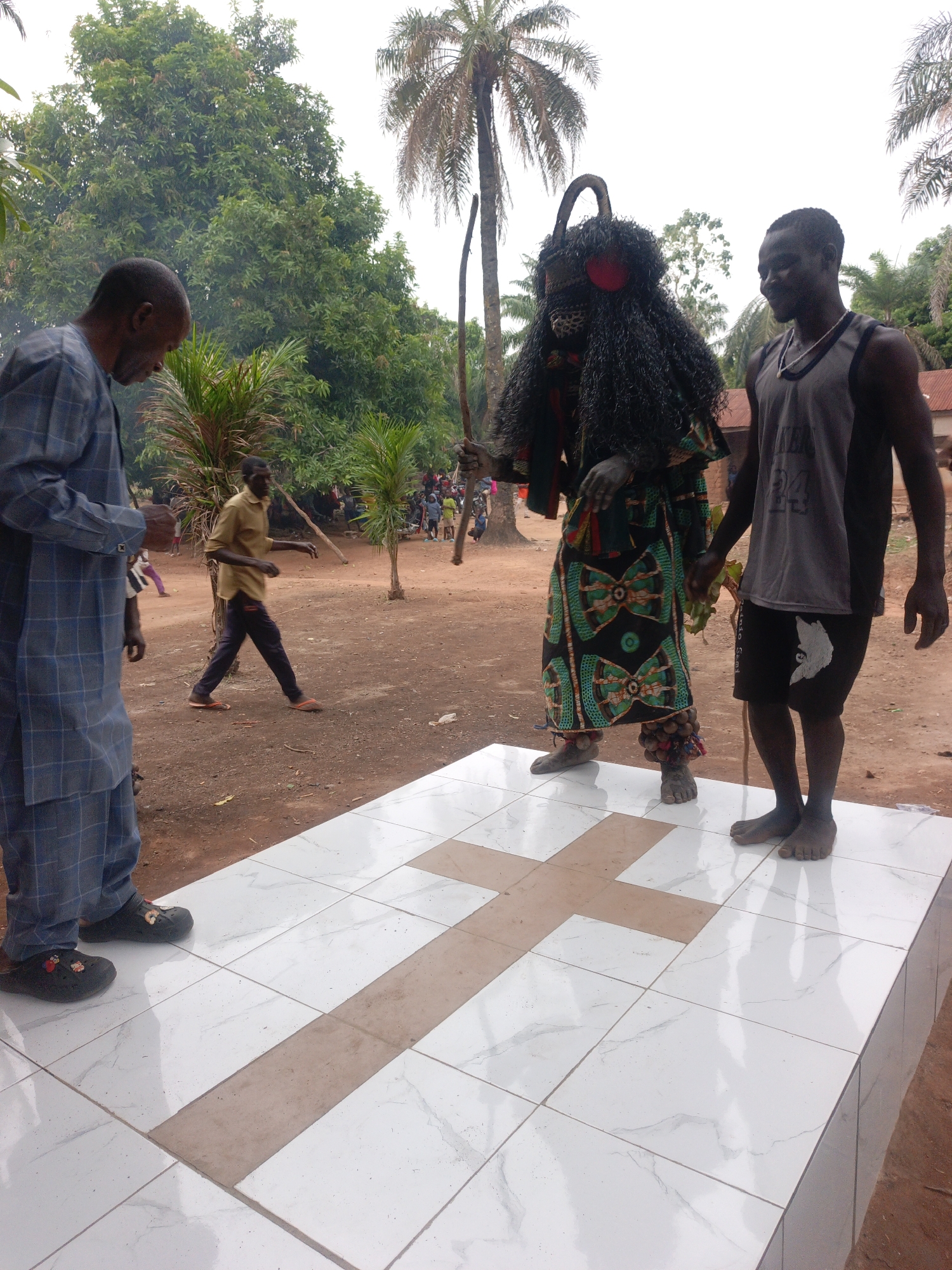 The image shows a ritualistic scene in what appears to be Akwanga, Nigeria. Centered is a person in a traditional costume with a large black fringed mask, horn-like protuberance and wrapped in colorful fabric with brown attachments on the ankles. To their left, an older man in a blue checkered suit looks down. To the right, a younger man in a grey tank top and black shorts stands barefoot, partially holding hands with the costumed figure. They all stand on a raised white tiled platform with a beige cross design. In the background, a figure in a yellow shirt walks past. A crowd of people are visible in the middle distance. The setting is an outdoor dirt lot with tropical trees and vegetation.
