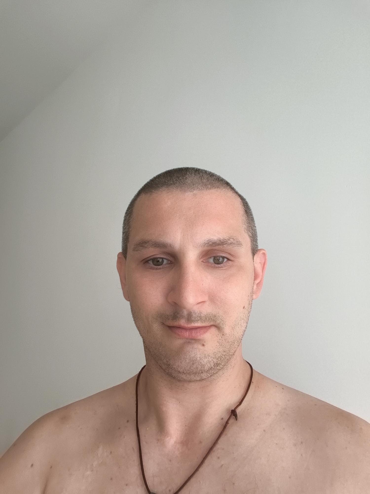 A close-up, head and shoulders shot of a man with a buzz cut, pale skin, and light brown eyes is centered against a plain, light grey wall. He is looking directly at the camera with a slight smile and his mouth closed. He has a bit of stubble on his chin and upper lip. A thin, dark brown cord necklace with a knot is visible around his neck and rests on his bare chest and upper shoulders. The man's skin shows some minor blemishes and marks. The environment appears to be indoors, with a subtle shadow cast on the wall in the upper left quadrant of the image. No specific location in Wolgast, Germany can be identified from the visual information provided.