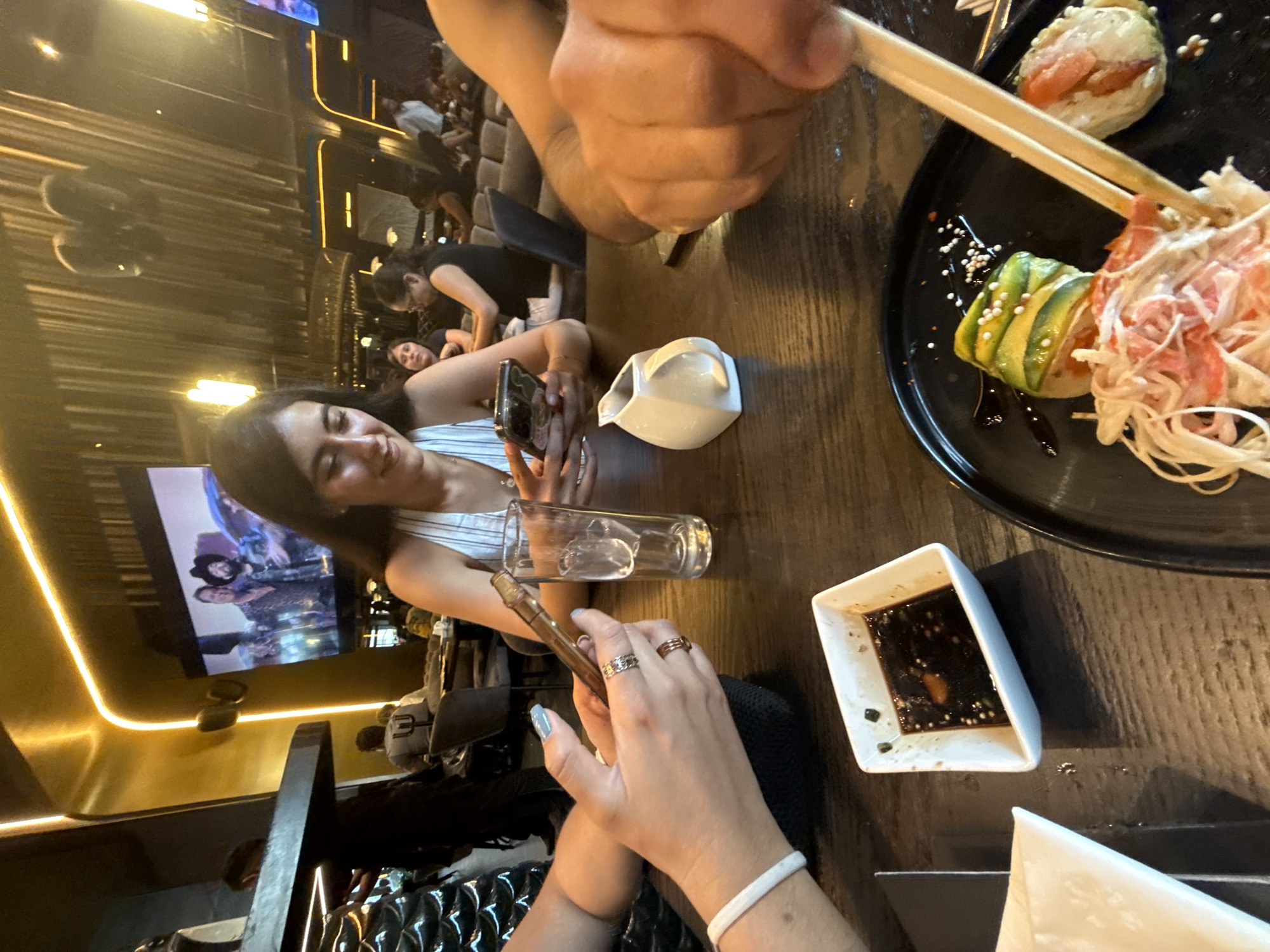 Captured indoors, the scene unfolds around a sushi meal. A woman sits at the table, casually holding her phone. A glass of liquid and a small pitcher sit nearby. In the foreground, chopsticks hold a piece of sushi. A small dish containing dark liquid and a folded napkin are visible. The background includes other people and a television. This photo appears to have been taken in San Bernardino Tlaxcalancingo, Mexico.
