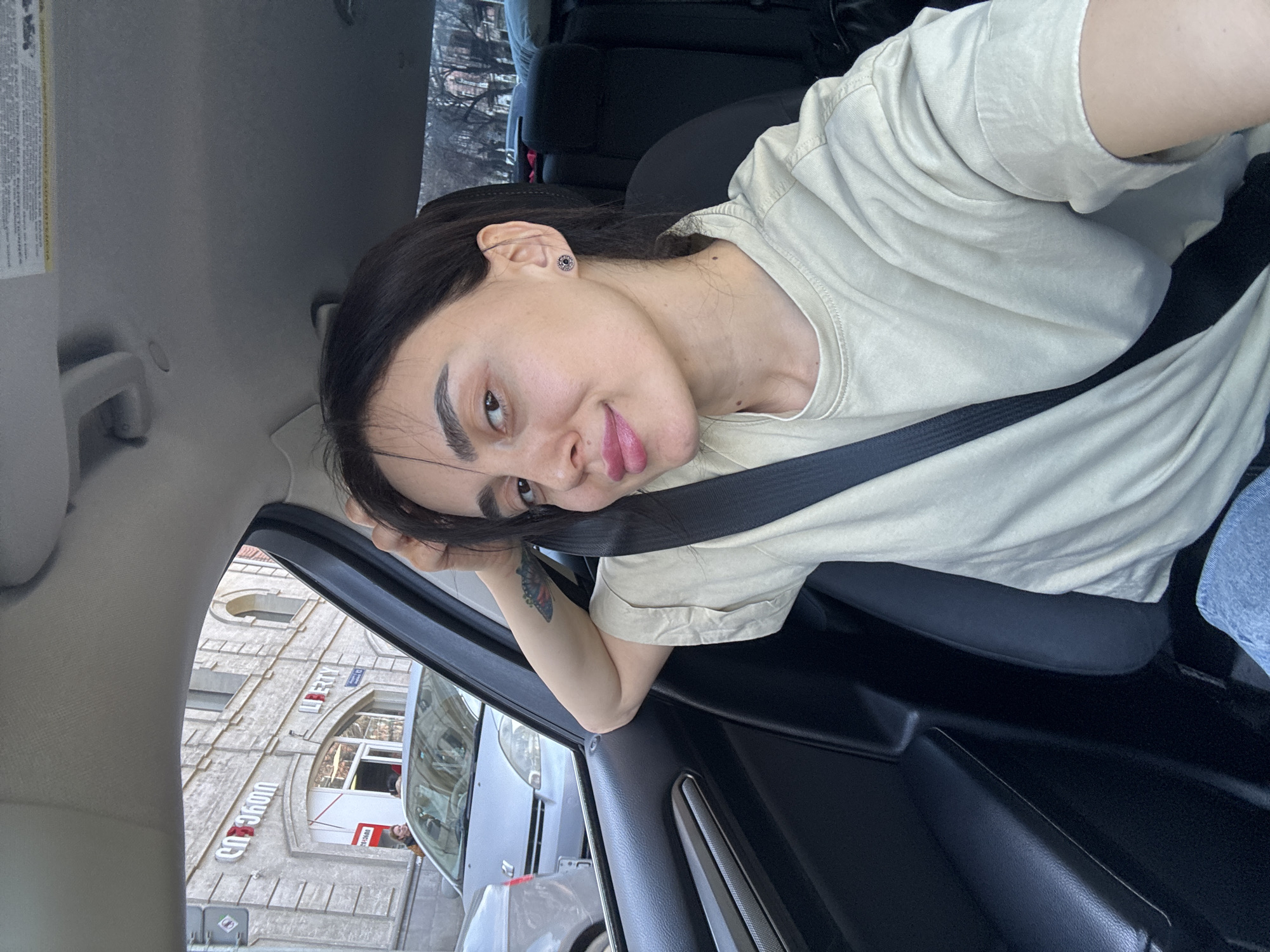 A woman wearing a beige t-shirt and a seatbelt is in a car, possibly in Tbilisi, Georgia. The photo appears to be a selfie, and the woman is leaning her head to the side. Outside the car window, there is a building with the Russian or Georgian word "СОЮЗ" (Union) written on it, and further up, "LIBERTY" is visible.