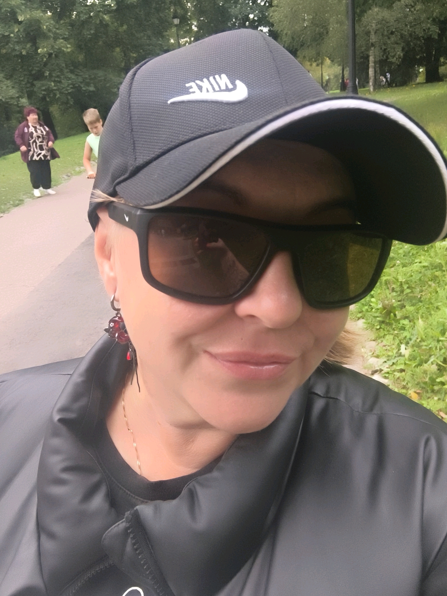 A medium shot selfie shows a woman from the chest up, wearing a black Nike baseball cap and dark sunglasses. She has blonde hair and is wearing a black puffer jacket over a black shirt. She has on red cherry-shaped earrings and a delicate gold necklace. In the background, two figures are visible on a paved path in a park. To the left, an older woman with dark hair is wearing a patterned shirt and black pants. To the right, a young boy is on a scooter. The park setting includes green grass and trees. The image was taken in Moscow, Russia.