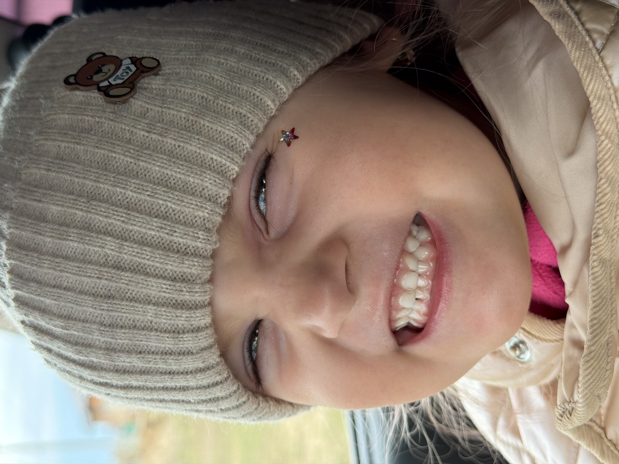 A young child with fair skin, wearing a light-colored knitted hat adorned with a teddy bear patch and a small star sticker near their eyebrow, is smiling widely. The child is also wearing a beige jacket with a pink undershirt. The background is blurred, possibly suggesting an outdoor location. There is no indication in the image that indicates that the picture was taken in Chaikovsky, Russia.