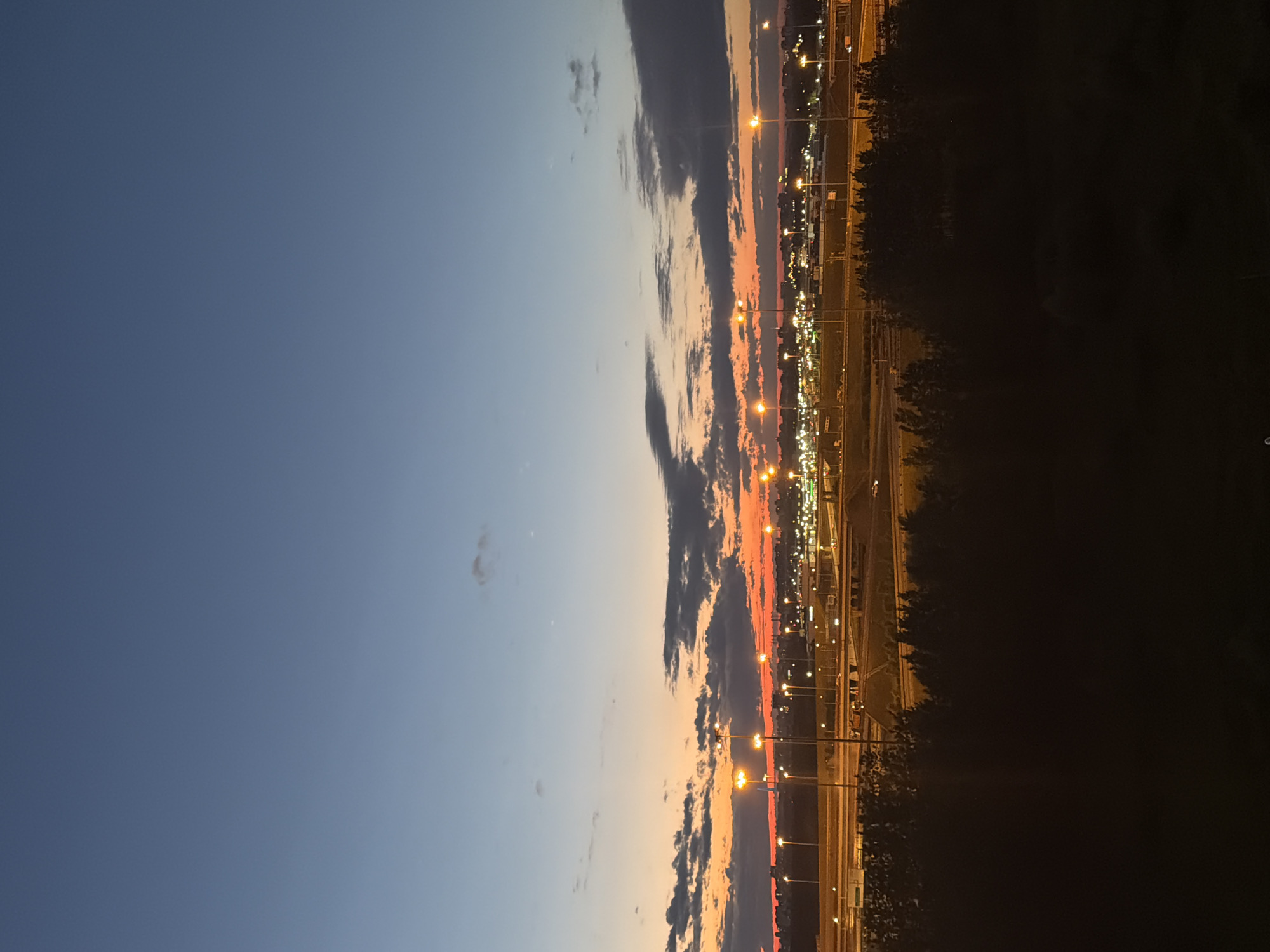 A wide-angle, vertical shot captures a twilight scene over a city, with the sky transitioning from a deep blue at the top to a vibrant orange and red near the horizon. Several scattered clouds, dark grey and tinged with orange from the setting sun, are visible in the middle ground. Below the sky, a line of tall streetlights with glowing orange lamps illuminate a highway or a complex road system. Beyond the road, a cityscape is visible, marked by numerous bright, yellow lights. In the foreground, the silhouette of dark trees creates a stark contrast against the illuminated background. The image appears to be taken from an elevated position, looking out over the urban landscape at dusk.