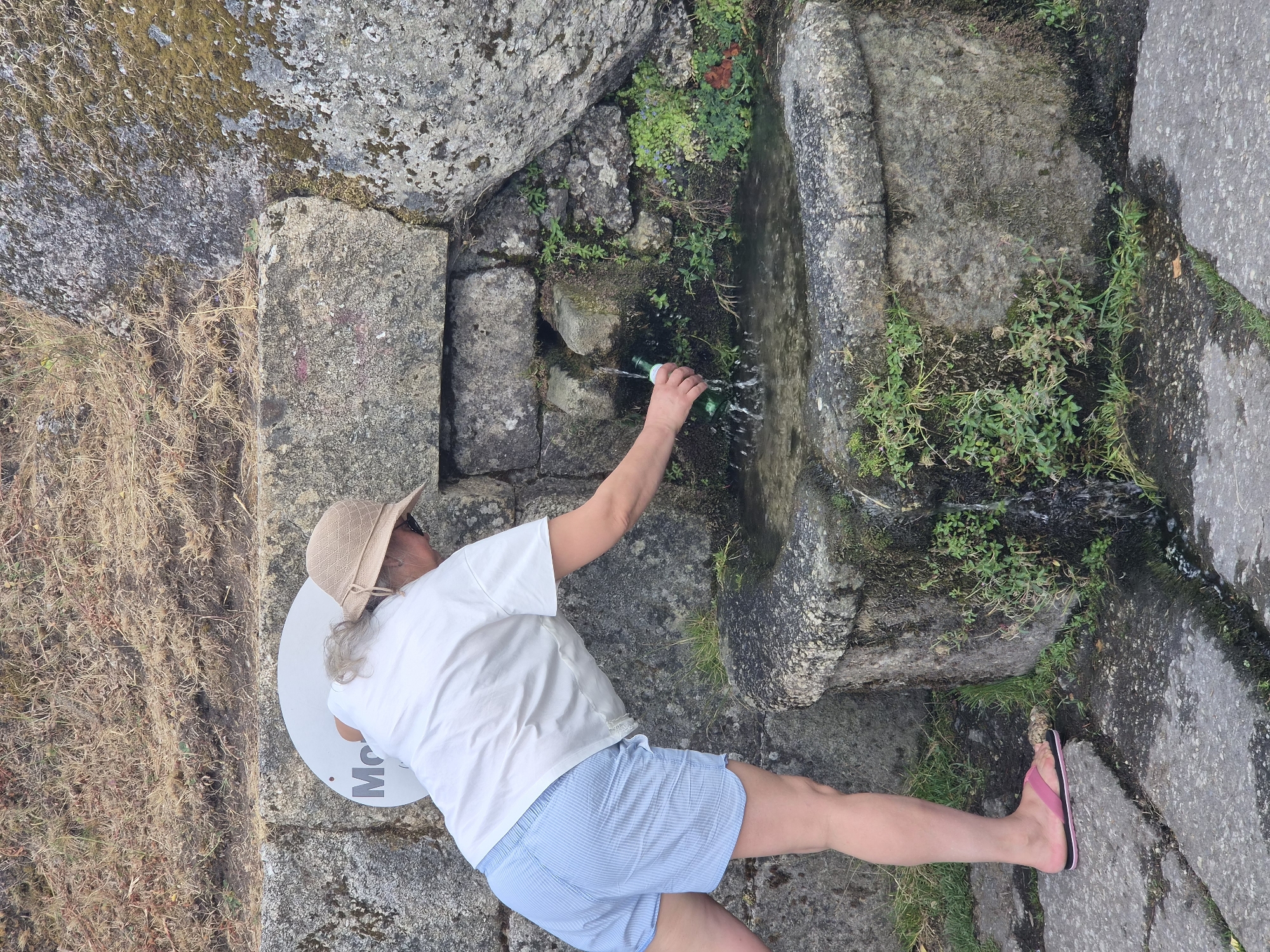 A person wearing a white t-shirt, light blue striped shorts, a tan sun hat, and pink flip-flops is filling a green bottle with water from a stone fountain. The fountain is built into a rocky, moss-covered structure with water trickling down. Green vegetation grows around the fountain and on the rocks. A circular white sign with the letter "M" and other unreadable text is visible behind the person. The ground is paved with large stone slabs. The scene appears to be outdoors, possibly in a rural or historical setting. The location is identified as a fountain in Gouveia, Portugal.