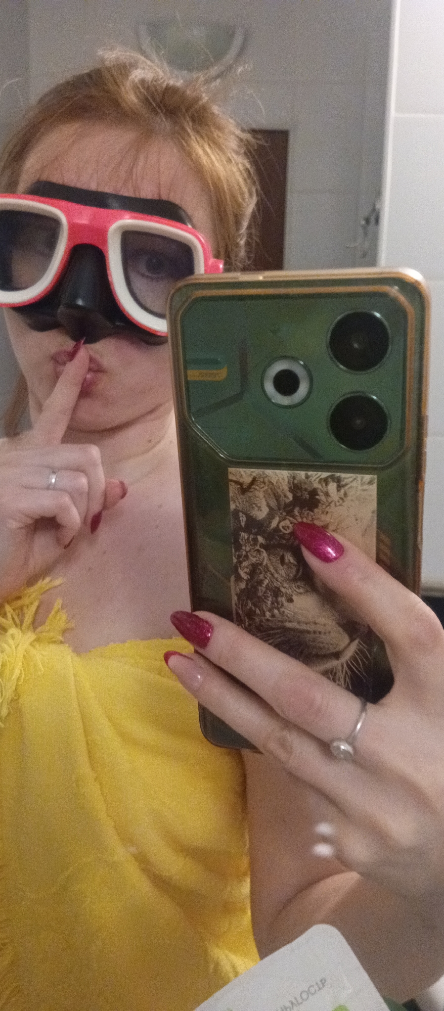 The image depicts a person holding a smartphone, possibly taking a selfie in a bathroom, judging by the tiles in the background. The person is wearing a scuba mask with a pink and white frame. She is wearing a yellow top and has red nail polish with white tips. She is holding her index finger to her lips in a shushing gesture. A silver ring is visible on both the ring finger of her left hand and the ring finger of her right hand. The smartphone has a dark green case with an image of a lion. The phone case also has three cameras on the back.