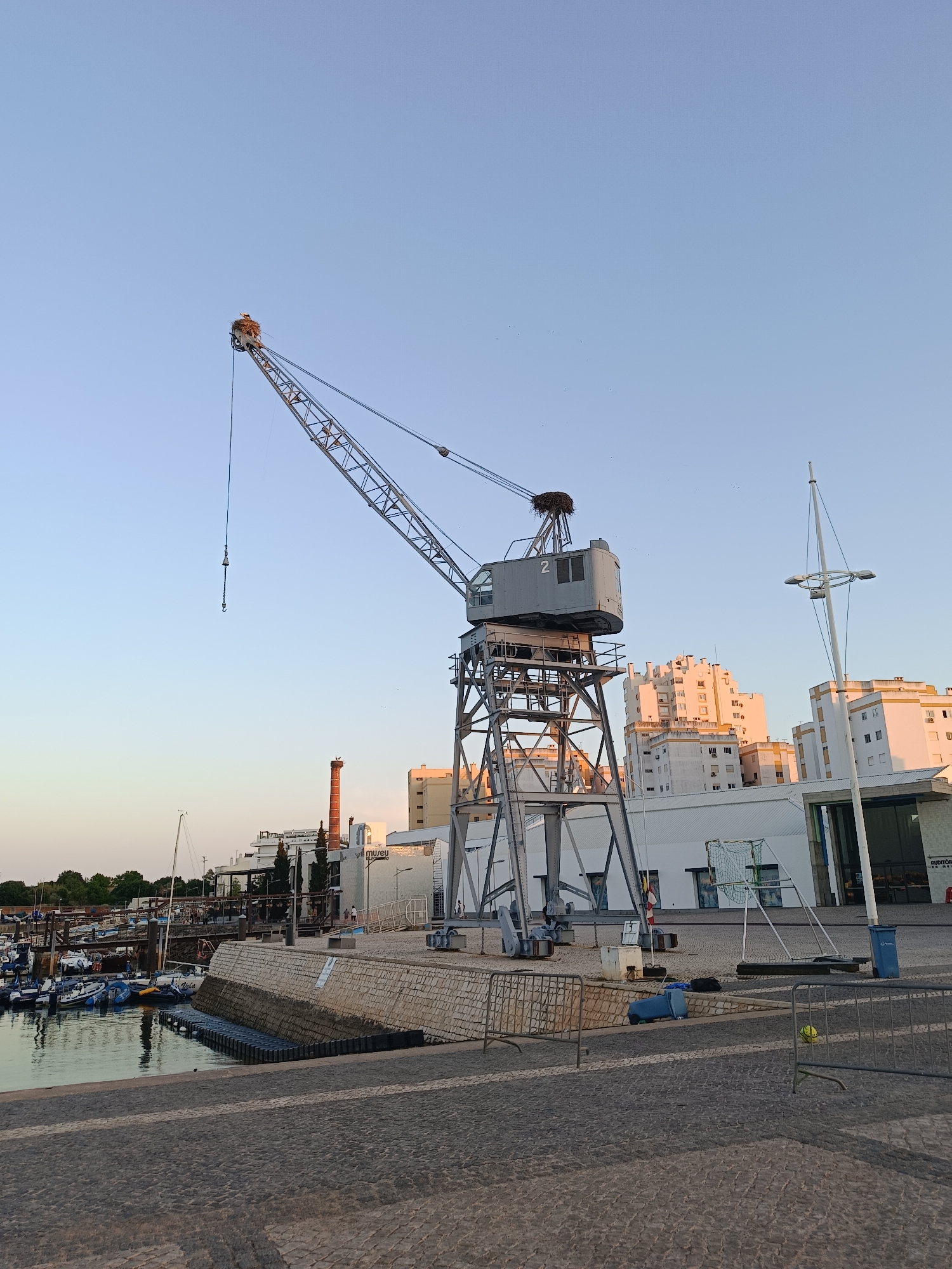 A wide-angle, full shot captures a harbor scene at dusk. In the foreground, a paved walkway leads towards a stone embankment bordering a body of water where several boats are docked. A large, grey industrial crane dominates the midground, its boom extending across the frame. A bird's nest is visible atop the crane's structure. Beyond the crane, a row of white buildings lines the waterfront, with a tall, reddish-brown chimney visible among them. The sky above is clear and pale blue, with a gradient towards a lighter hue near the horizon. The overall lighting suggests the late afternoon or early evening.