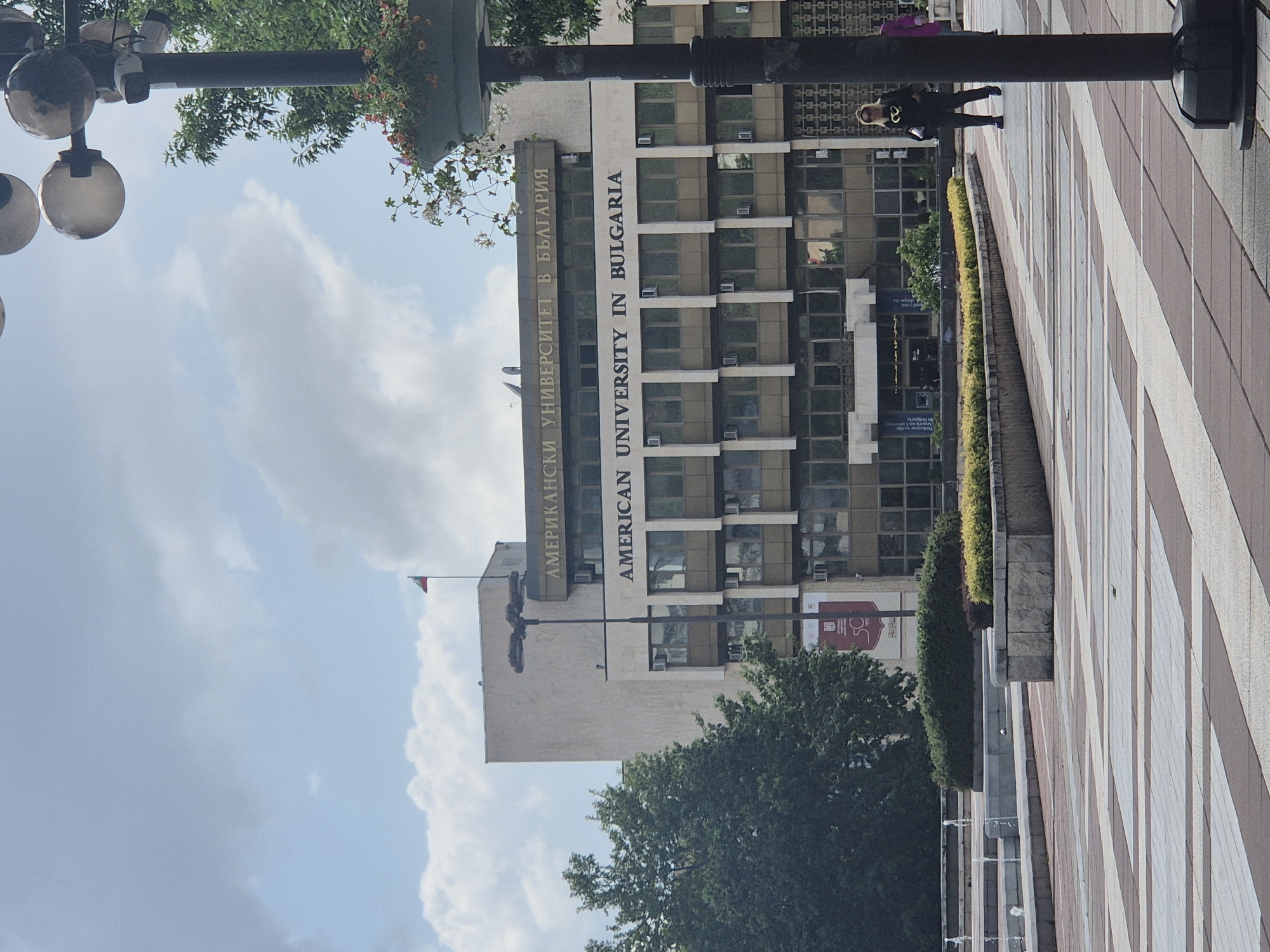 This image captures the exterior of the American University in Bulgaria, located in Blagoevgrad, under a partly cloudy daytime sky. The multi-story university building dominates the central and right portions of the frame, featuring a modern facade with numerous rectangular windows and visible air conditioning units.
Prominently displayed vertically on the building are the names of the institution in both Cyrillic and Latin scripts: "АМЕРИКАНСКИ УНИВЕРСИТЕТ В БЪЛГАРИЯ" and "AMERICAN UNIVERSITY IN BULGARIA." A small flag, possibly the Bulgarian flag or university flag, is visible at the top of the building. On the lower right side of the building, a red sign with a partially visible logo, possibly featuring the number "30," is attached.
In the foreground to the left, a large street light fixture with three spherical glass globes is visible, along with lush green foliage and what appears to be a hanging flower basket. To the right of the street light, a dark, thick pole stands.
The ground level features a wide, paved pedestrian area with alternating light and dark striped patterns, stretching towards the right side of the image. Along the edge of this paved area, near the university building, are neatly trimmed shrubs and planters. One person, dressed in dark clothing with blonde hair, is partially visible standing near the dark pole on the right side of the paved area, suggesting a typical day in an urban university setting. The overall atmosphere is bright and calm, indicative of good weather.