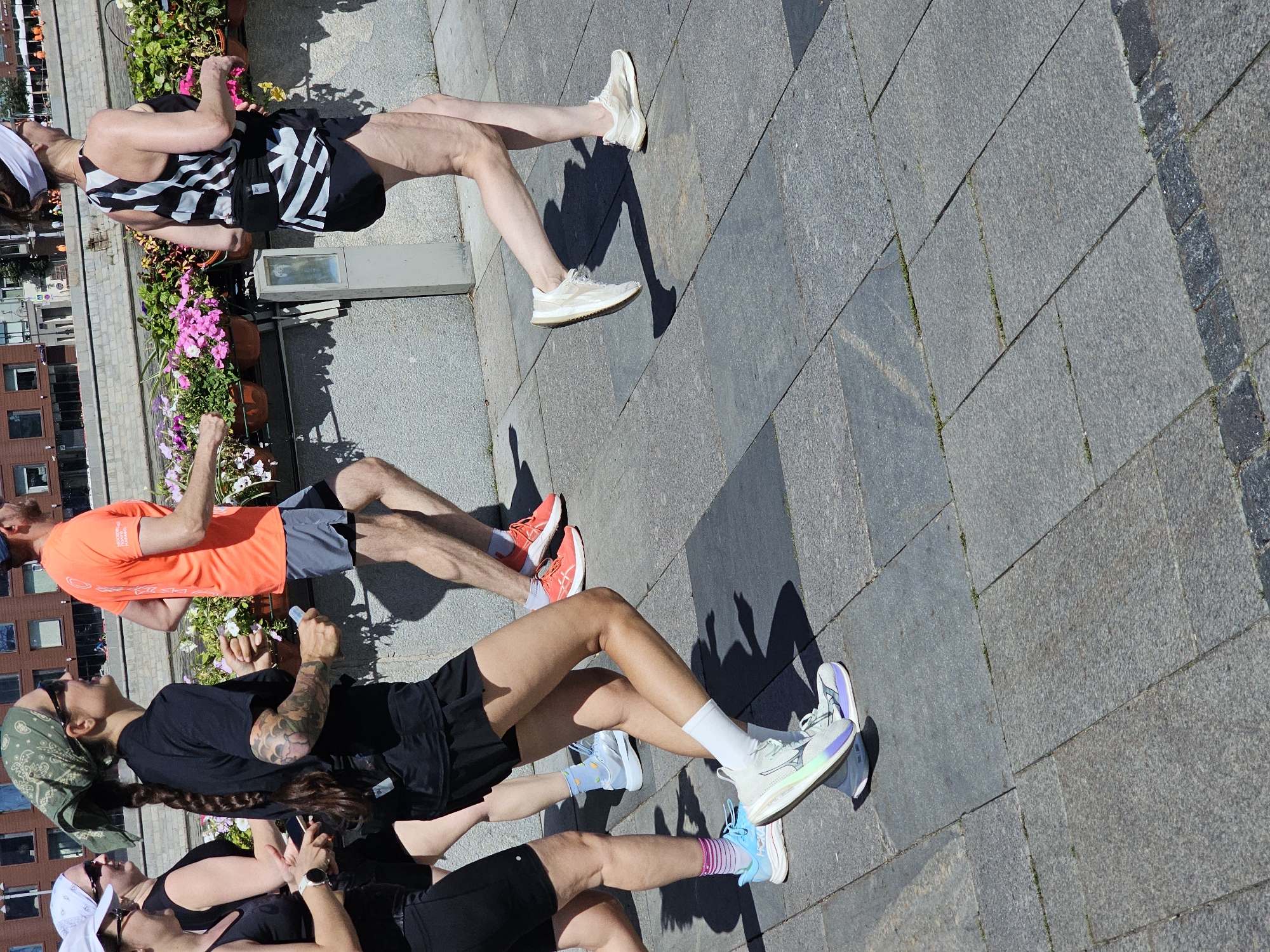Four individuals are running on a paved path alongside a body of water. The person in the foreground on the left wears a black tank top, black shorts, and light blue and white athletic shoes. Next to them, to the right, a person in a black t-shirt, black skirt, and white and light green athletic shoes with purple and blue accents is running. Behind them, a person in an orange t-shirt and gray shorts is running. To the far right, a person in a black and white patterned tank top and black shorts is running. All individuals appear to be participating in a race or training exercise. The environment features a stone wall and planters filled with pink and white flowers. The path is made of large, square stone pavers.