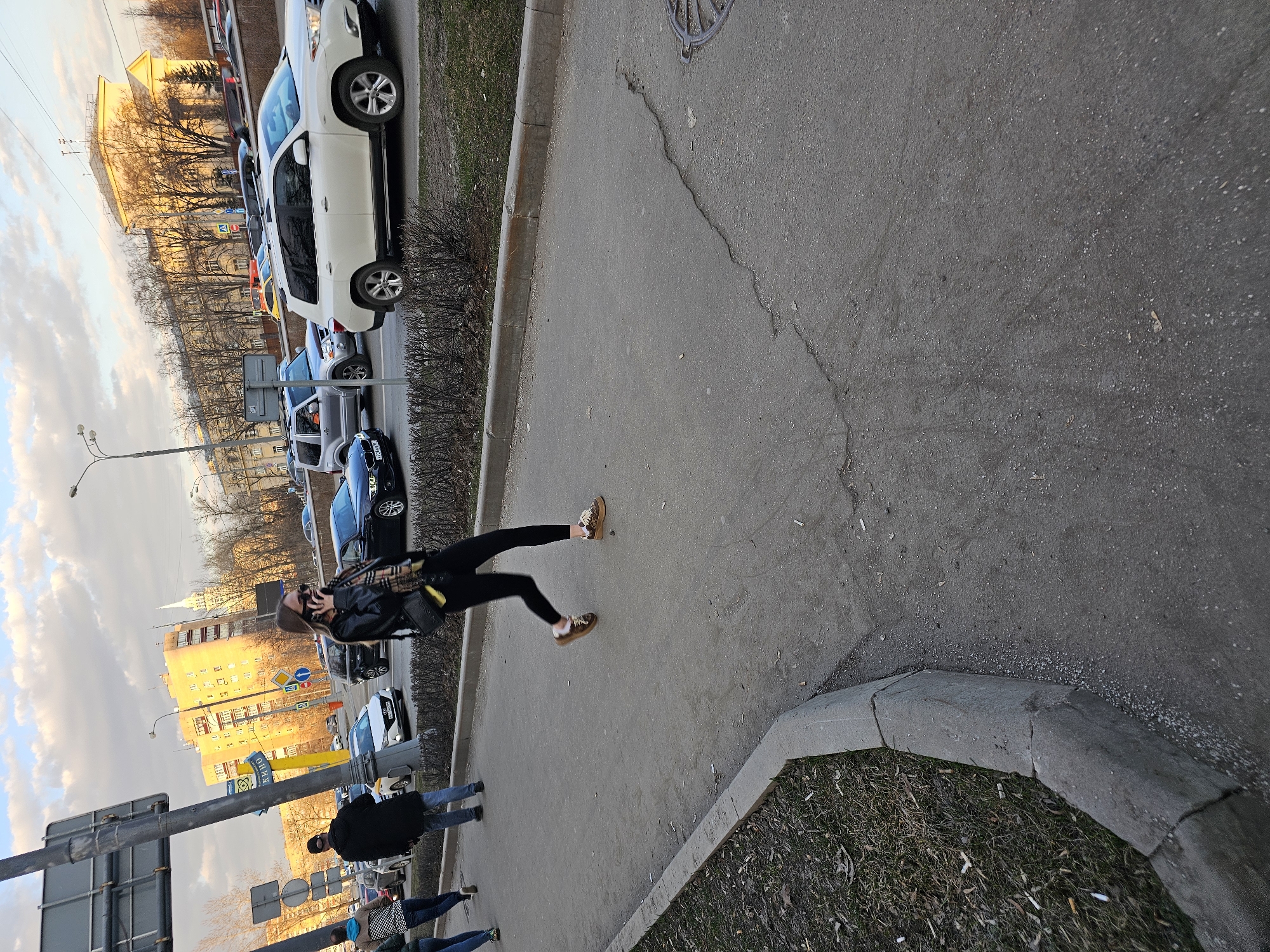 The image depicts an urban street scene in Khimki, Russia, specifically on Leningradskaya Street, evidenced by the prominent "КИНО" (KINO) sign, indicating proximity to the Rodina Cinema, which is the large yellow building in the background.
In the foreground, an asphalt pedestrian sidewalk runs parallel to a multi-lane road. The sidewalk exhibits multiple cracks, particularly towards the right side. A concrete curb separates the sidewalk from a narrow strip of ground cover containing sparse dry vegetation, fallen leaves, and numerous discarded cigarette butts. A circular metal manhole cover is partially visible on the far right of the sidewalk.
Three individuals are present on the sidewalk. A female in the mid-foreground is captured mid-stride, moving towards the right side of the frame. She is engaged in a phone conversation, holding a mobile phone to her right ear. She wears dark sunglasses, a black leather jacket, dark leggings, and light brown low-top shoes with light-colored accents. A patterned scarf and a black shoulder bag are also visible. To her left, in the mid-background, a male figure wearing a dark coat, blue jeans, and a dark baseball cap walks towards the left side of the frame. A third individual, partially obscured on the far left, is wearing a blue hooded top and jeans, walking away from the camera.
The multi-lane asphalt road is populated with various passenger vehicles, suggesting traffic congestion. Prominent vehicles include a white Toyota Highlander SUV in the mid-ground right and a silver Mitsubishi Pajero/Montero in the mid-ground left. A black sedan (possibly BMW) and a white sedan are also visible among other vehicles.
On the left side of the frame, a large grey utility pole with attached signs, including the yellow "КИНО" sign with blue Cyrillic text, stands prominently. In the background, the large multi-story yellow Rodina Cinema building is illuminated by sunlight. To its right, a white spire of a building is visible. Various other urban buildings, streetlights, traffic signs, and bare deciduous trees line the street. The sky above is partially cloudy, with patches of blue and golden light. The scene appears to be set during late afternoon or early evening.