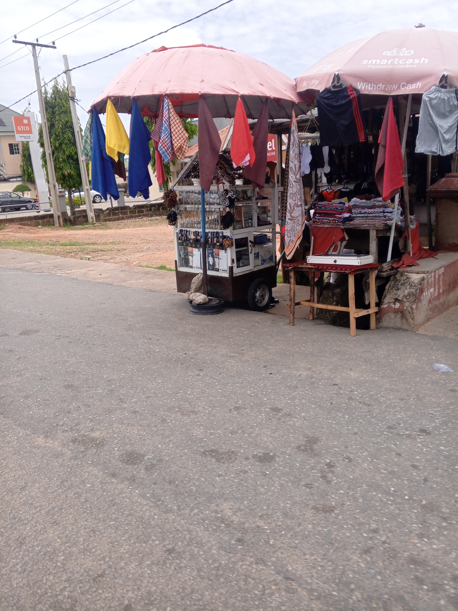 The image depicts a street scene in Jalingo, Nigeria, with a focus on two stalls displaying clothing and accessories under large umbrellas. The stall on the left features a variety of colorful fabrics hanging from an open red umbrella, along with smaller items like jewelry displayed on a rack. This stall appears to be a mobile cart with wheels. To the right, another stall operates under a larger, tan umbrella, which has "Smartcash" and "Withdraw Cash" printed on it. This stall displays shorts and other clothing items hanging, as well as stacks of folded fabrics. A small wooden table in front of this stall holds a white object, possibly a cash register or a portable scale. In the background, a GTCO (Guaranty Trust Holding Company) sign is visible on a building, and a few vehicles are parked along the roadside. The overall environment suggests a commercial street, likely a market area, with an overcast sky.