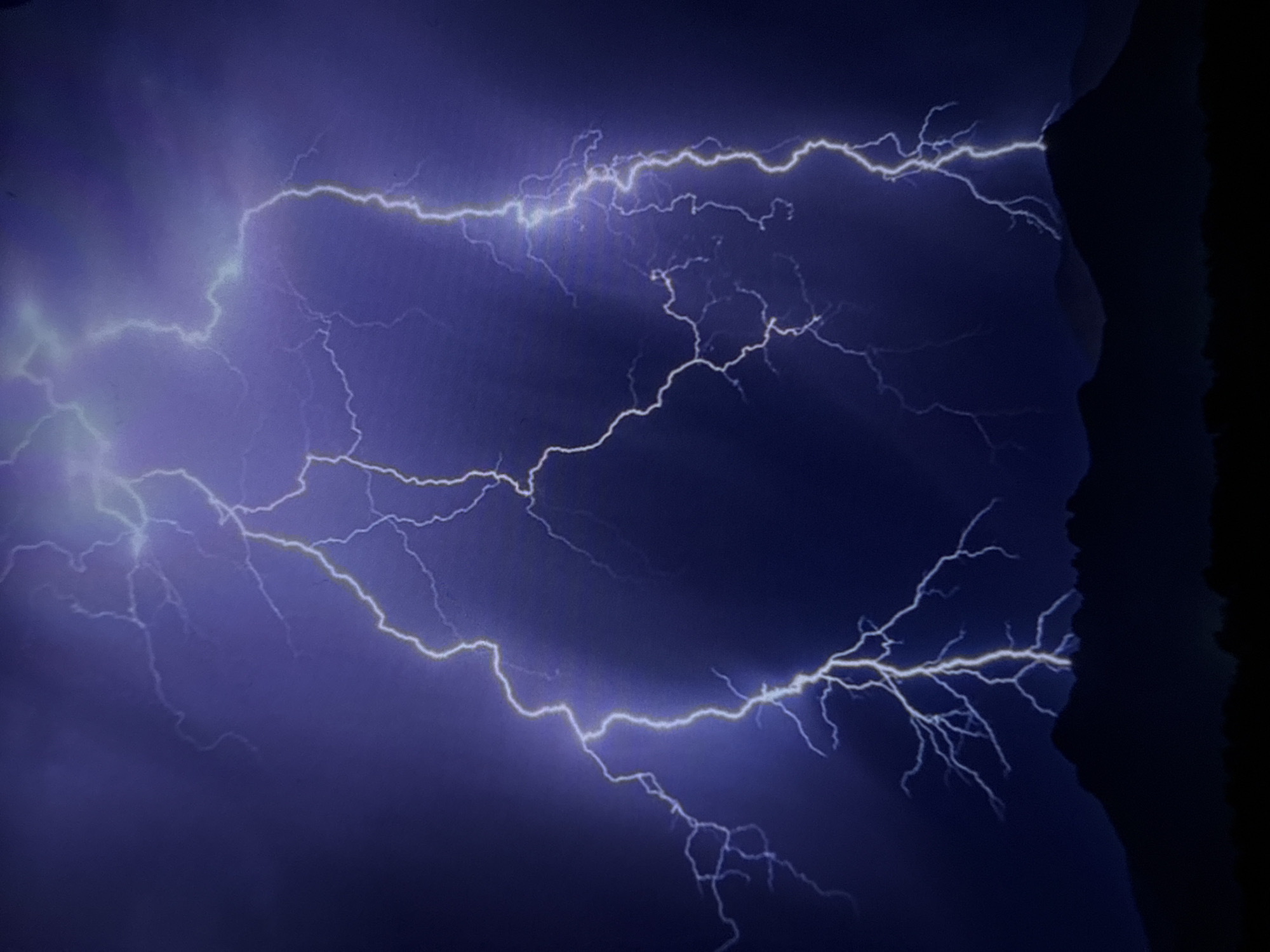 A nighttime photograph depicts a dramatic lightning storm over a silhouetted mountain range. Multiple bolts of brilliant white and blue lightning illuminate the dark, turbulent sky, casting an ethereal glow. The jagged lines of the lightning strike downward, some appearing to hit the peaks of the mountains. The foreground shows the dark, undulating shapes of mountains and a slightly lighter, flatter silhouette of distant hills or vegetation. The sky is a deep, ominous purple-blue, with wisps of clouds adding texture. There are no visible people, structures, or signs of human activity other than what might be implied by the landscape. This scene does not provide any specific indicators of location within the city of Varna, Bulgaria.