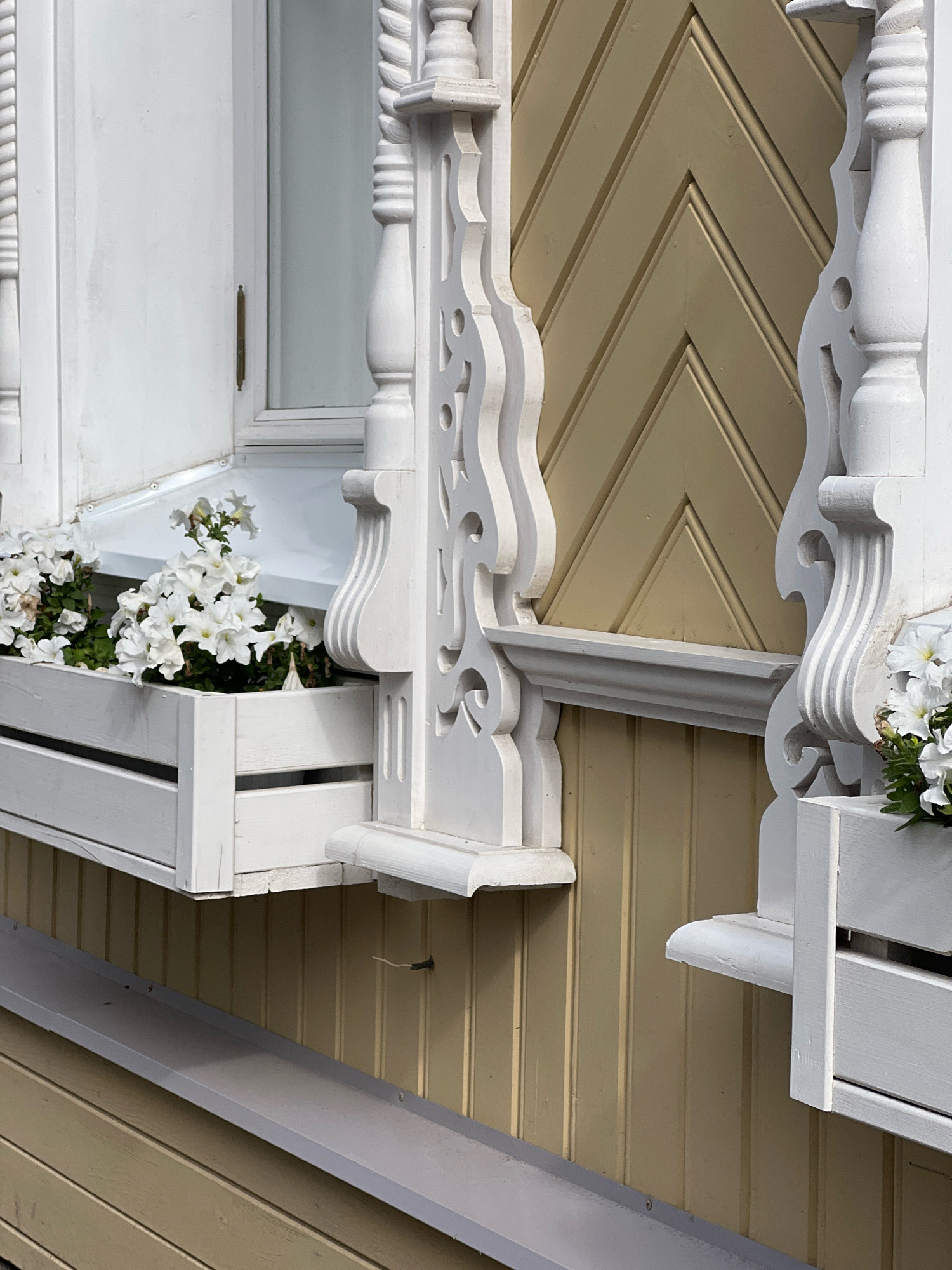 A close-up, eye-level, outdoor shot captures the exterior of a light yellow wooden house with white decorative trim and window boxes. The wall is clad in vertical wooden planks, with a diagonal pattern of wooden siding above the window. White petunias are planted in a white wooden window box on the left, and another partially visible window box is on the right. Ornate white wooden carvings adorn the area around the window. A gray metal gutter runs along the bottom of the image. The scene is well-lit, suggesting daytime.