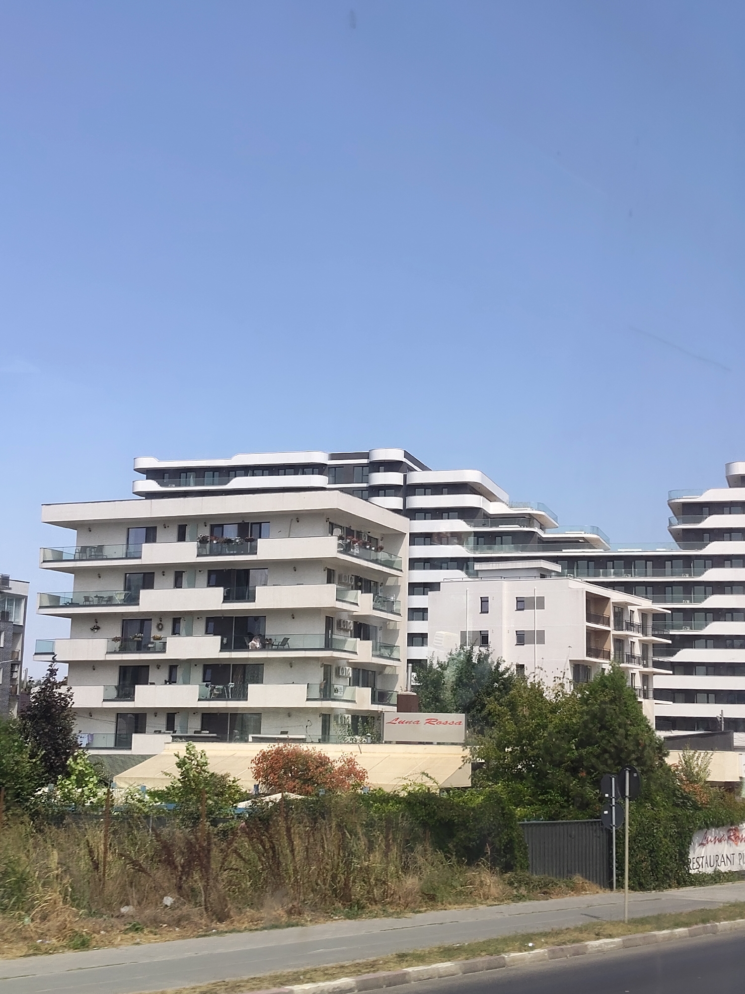 The image shows a modern residential building complex with a clear blue sky in the background. The buildings are multi-story with white facades and balconies with glass railings. There are several levels of staggered balconies, creating a dynamic architectural design. Some balconies have outdoor furniture, and plants are visible on some of them. In the foreground, there is overgrown vegetation, including tall grasses and shrubs, and a paved road or sidewalk runs along the bottom of the frame. A fence is visible behind the vegetation. A sign with "Luna Rossa" and "RESTAURANT PIZZA" is partially visible on the right side of the image. The overall scene depicts an urban environment with contemporary architecture and a touch of greenery.